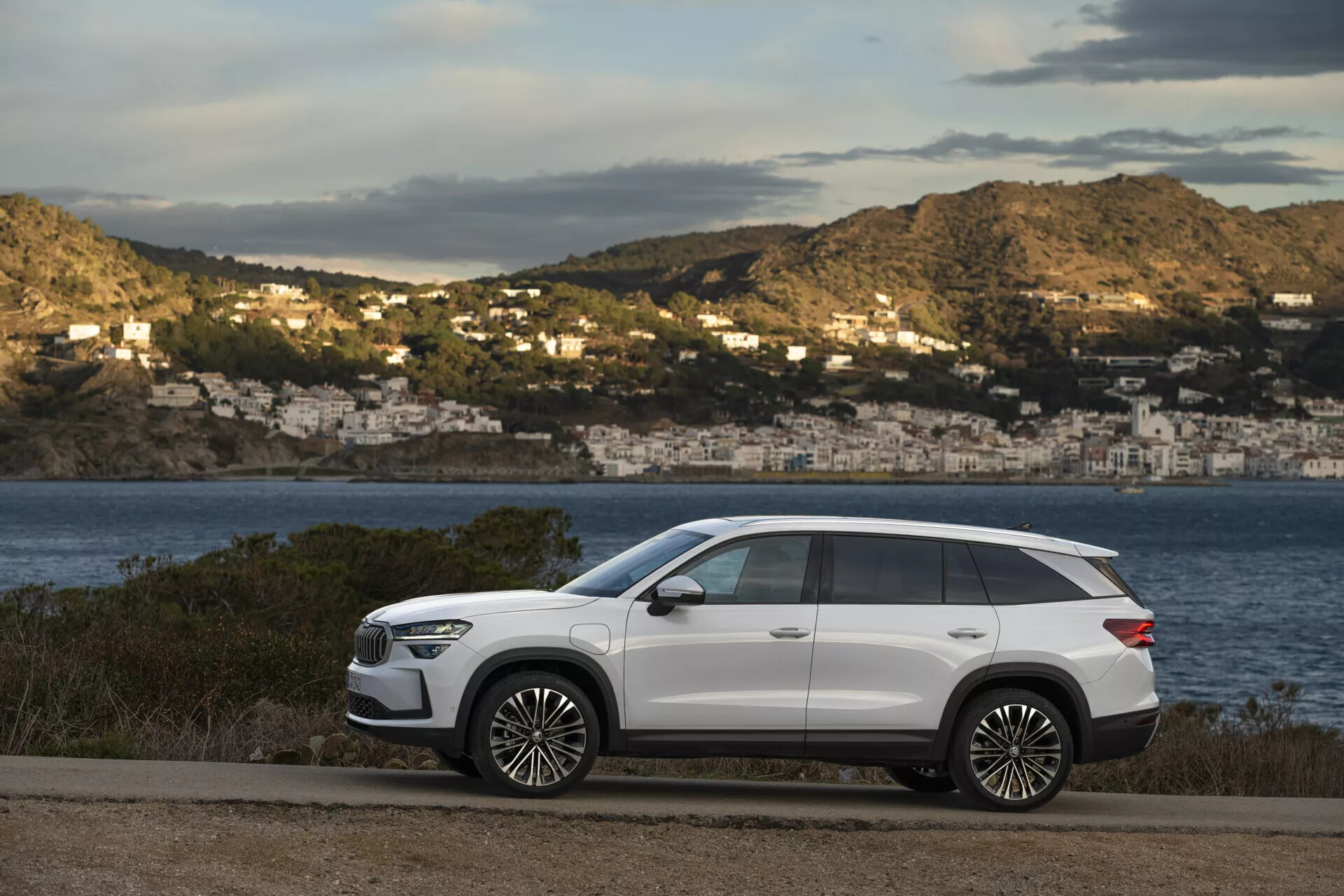 SUV moderno Skoda Kodiaq 2024 estacionado numa estrada costeira com uma vista cênica de uma baía e casas de encosta ao fundo, apresentando um design elegante, distância entre eixos longa, alta altura ao solo, linhas de carroceria nítidas, puxadores de porta embutidos, vidros traseiros tintados, barras de teto, grandes rodas de liga leve, faróis LED, espelhos laterais com sinais de direção, e teto solar panorâmico, tudo contra um acabamento exterior branco contrastante.