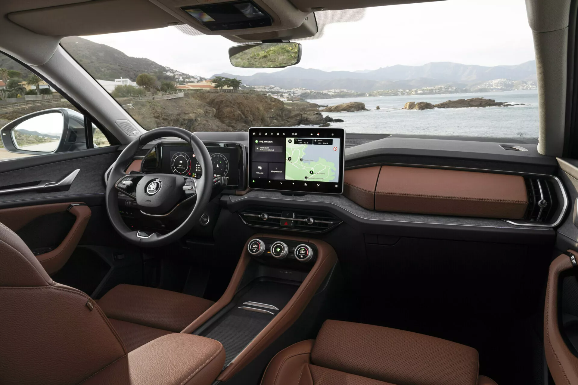 Vista interior do Skoda Kodiaq 2024 da perspectiva do passageiro mostrando o painel, volante multifuncional com logotipo, cluster de instrumentos digitais, grande sistema de infotainment com tela sensível ao toque e GPS, assentos de couro marrom luxuosos e design elegante do console central, contra uma paisagem costeira serena fora das janelas