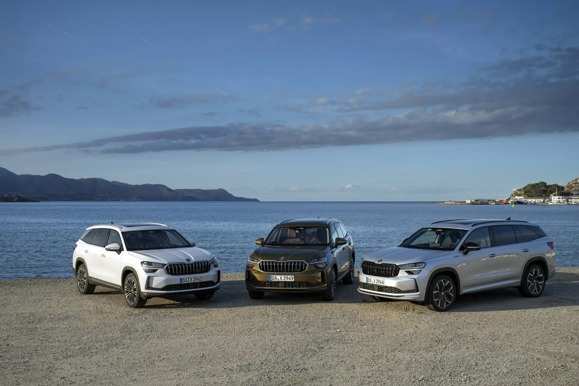 Linha de SUVs Skoda Kodiaq 2024 em cores branca e escura, vista lateral frontal, estacionados em uma orla com montanhas e um céu parcialmente nublado ao fundo