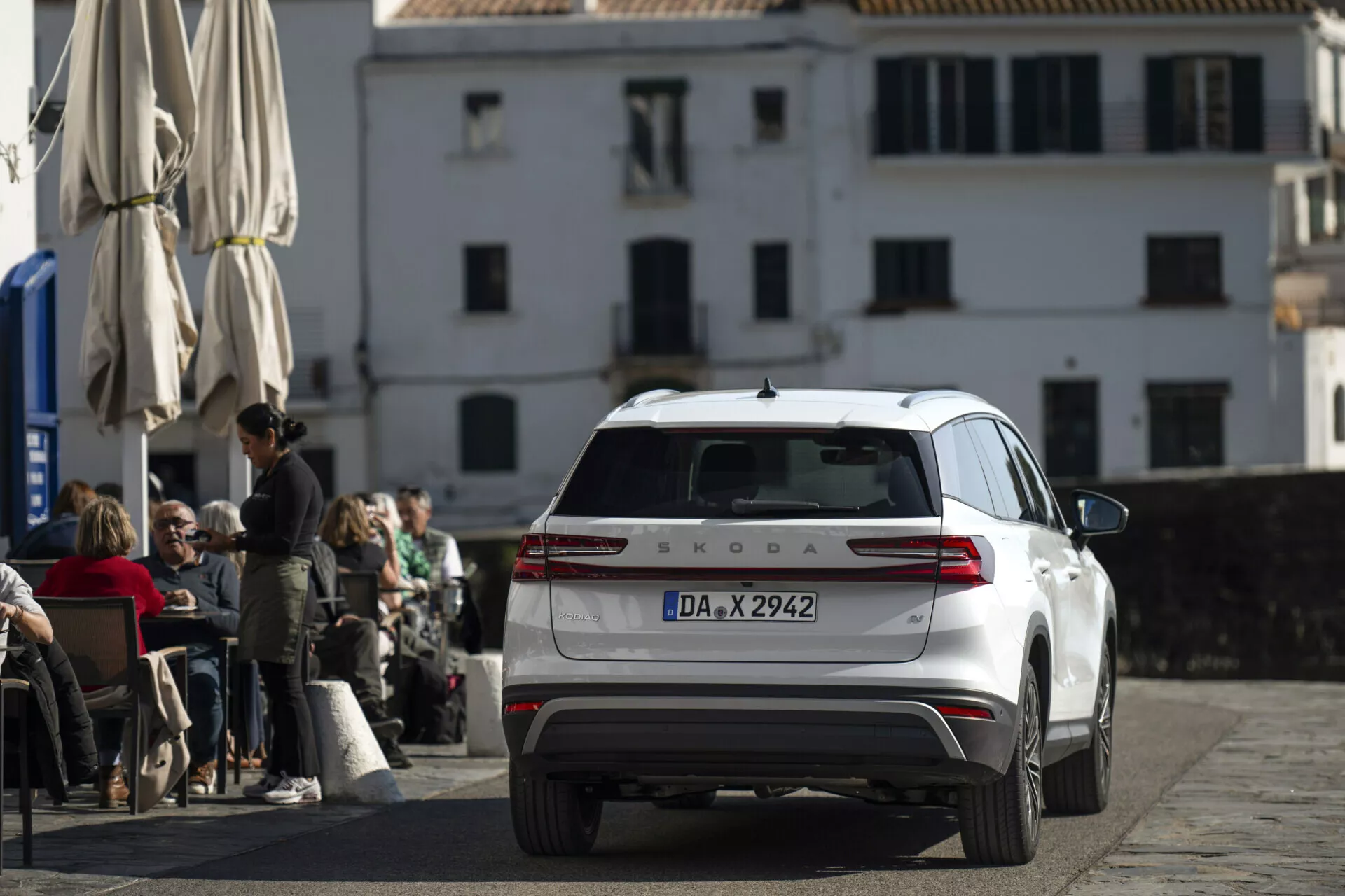 Škoda Kodiaq branco de 2024 fotografado de um ângulo traseiro e ligeiramente elevado, exibindo a parte traseira completa, lanternas traseiras, tampa do porta-malas, janela traseira com limpador e teto com antena e barras, estacionado em uma rua de paralelepípedos em frente a um café com arquitetura europeia clássica ao fundo.