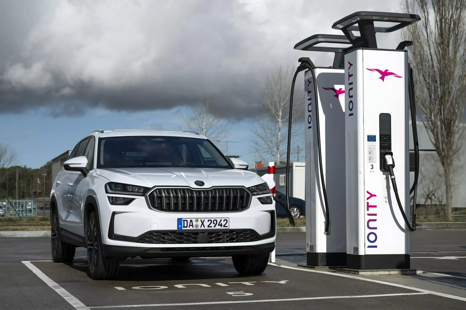 SUV branco Skoda Kodiaq 2024 estacionado numa estação de carregamento eléctrico, apresentando a vista do quarto frontal com faróis LED e luzes diurnas, enfatizando suas capacidades elétricas ou híbridas e design de grade luxuoso.