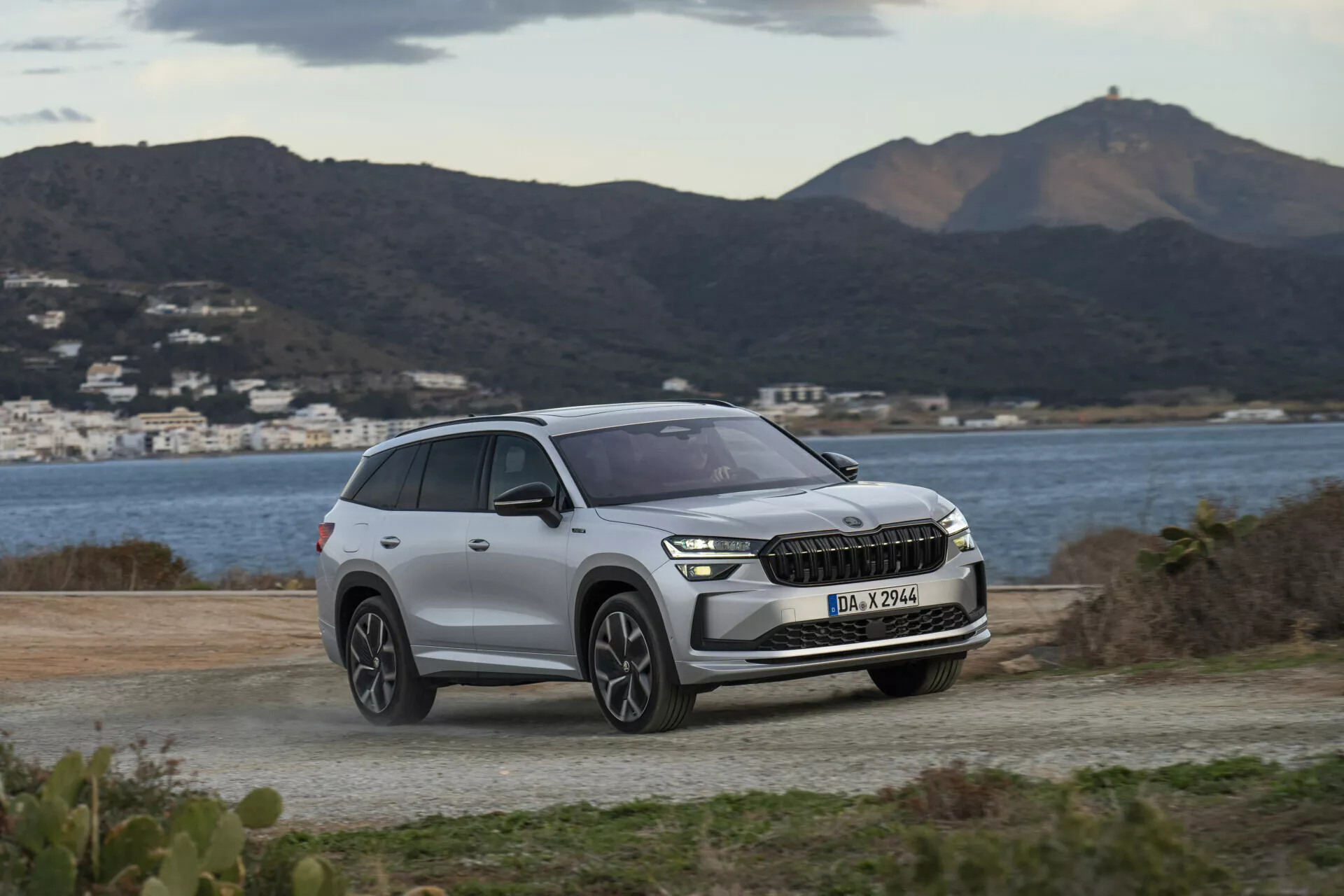 SUV Skoda Kodiaq 2024 estacionado em uma superfície não pavimentada, com um cenário de colinas e água ao fundo, destacando os faróis modernos em LED do veículo, grade frontal com possível integração ADAS, e design contemporâneo das rodas sob uma luz diurna suave e nublada.