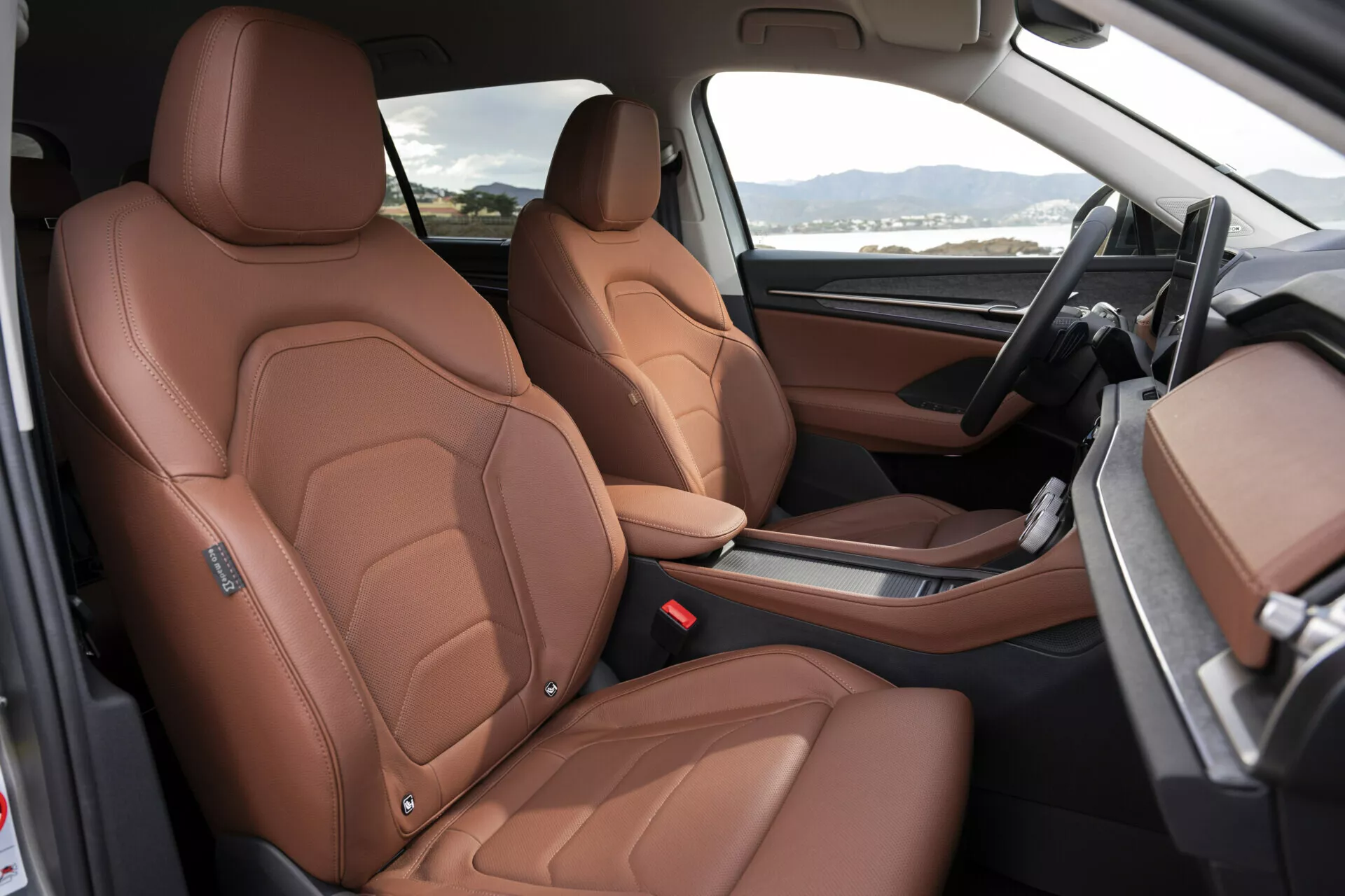 Vista interior do Škoda Kodiaq 2024 exibindo assentos dianteiros em couro marrom luxuoso com costura detalhada, ajuste elétrico dos assentos e uma visão dos elementos de design de alta gama como os reforços laterais inspirados no desporto, painel de bordo bicolores preto e marrom, e consola central minimalista com alavanca de câmbio automática, do ponto de vista da porta do lado do passageiro aberta