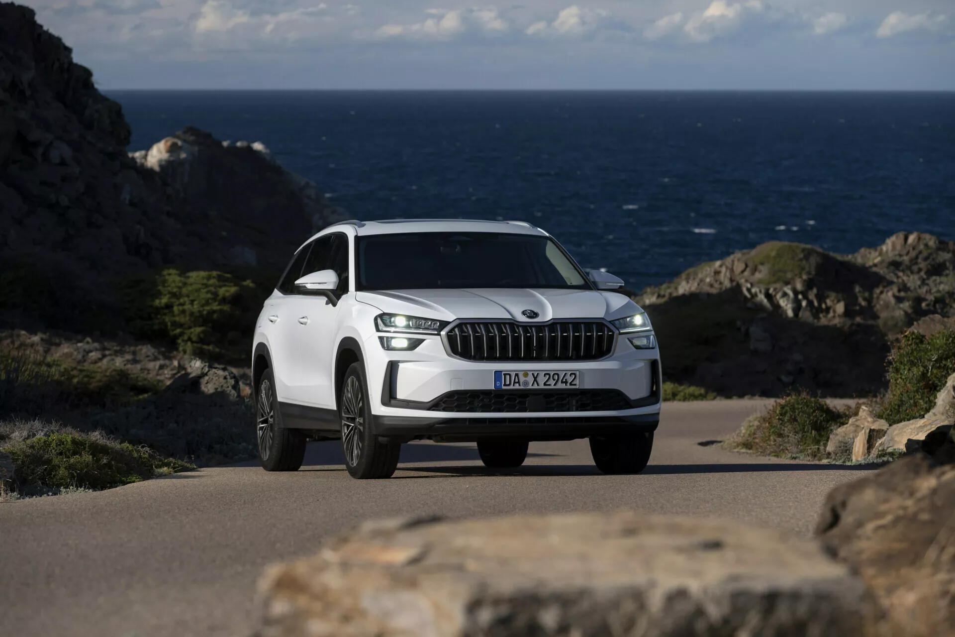 SUV branco Skoda Kodiaq 2024 estacionado em uma estrada costeira com o oceano ao fundo, apresentando uma grade frontal proeminente com ripas verticais cromadas, luzes diurnas de LED, rodas de liga leve multirraios e uma visão detalhada de três quartos da frente