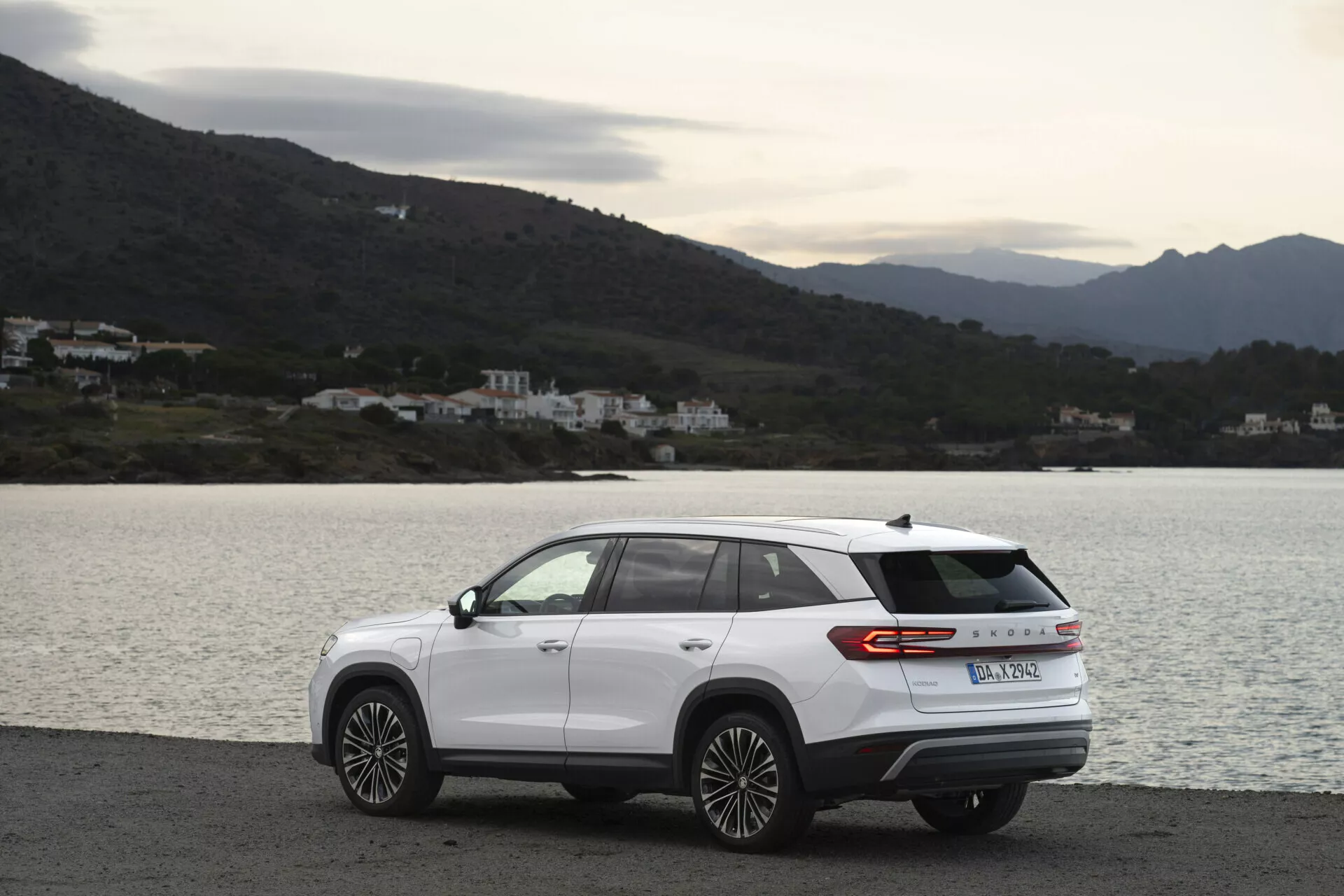 SUV moderno Skoda Kodiaq 2024 estacionado de lado em uma estrada costeira cênica, apresentando design aerodinâmico, faixa de luz traseira e rodas de liga leve de multi-raios, em um dia nublado com iluminação natural difusa, com um fundo de água, colinas e edifícios