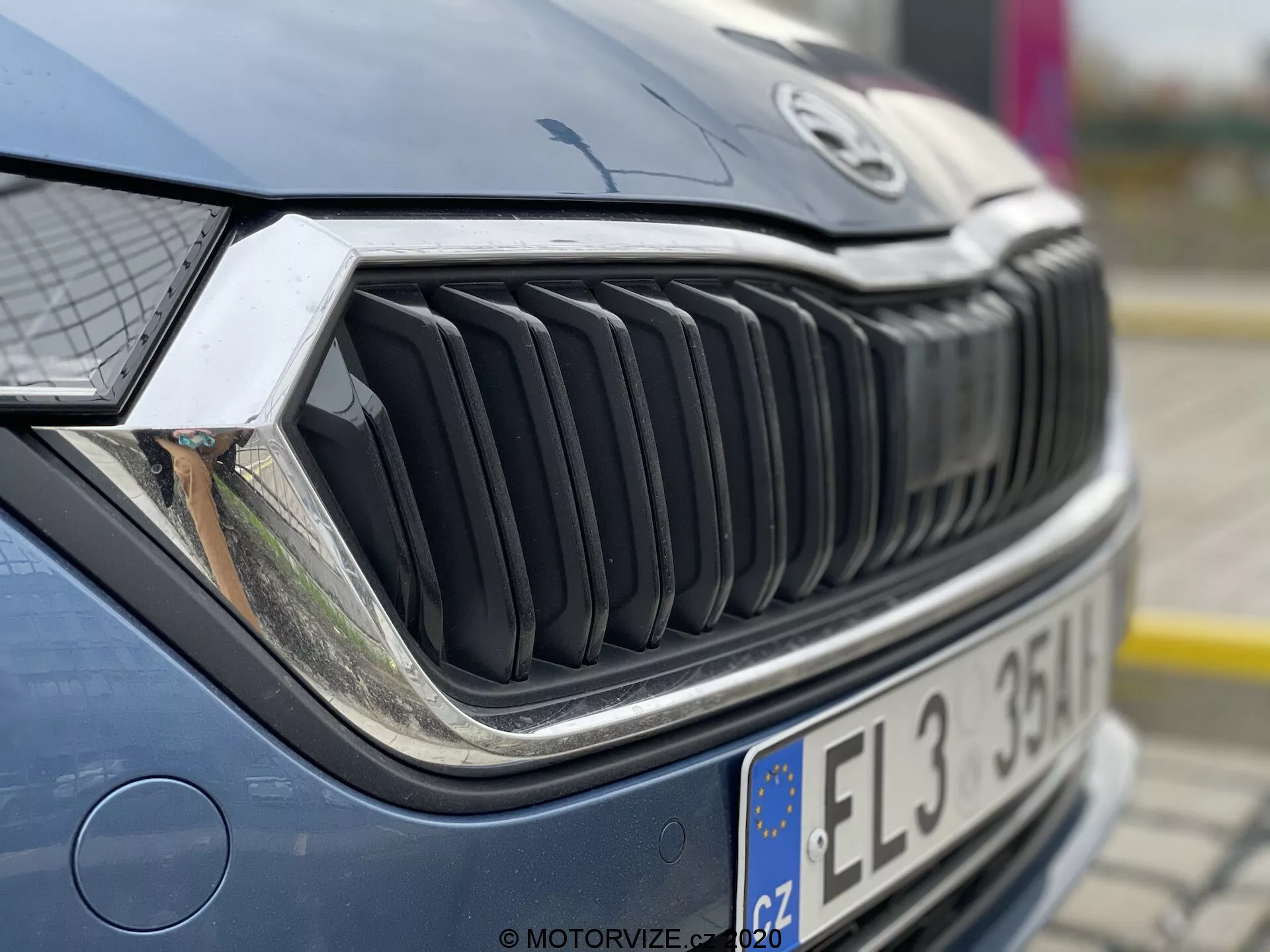 Imagem em close-up da seção frontal de um carro, focando na grade e no farol. Tirada de um ângulo dianteiro direito ligeiramente mirando para cima, a foto mostra uma visão detalhada do lado direito da grade, do farol direito e de parte do capô. As lâminas verticais da grade são proeminentes, indicando um design arrojado. O farol complexo sugere tecnologia de iluminação avançada, como LED ou sistemas adaptativos. O capô está ligeiramente desfocado, destacando a textura da grade e do farol. O carro está bem conservado, com reflexos indicando um cenário ao ar livre em um dia nublado ou à sombra.