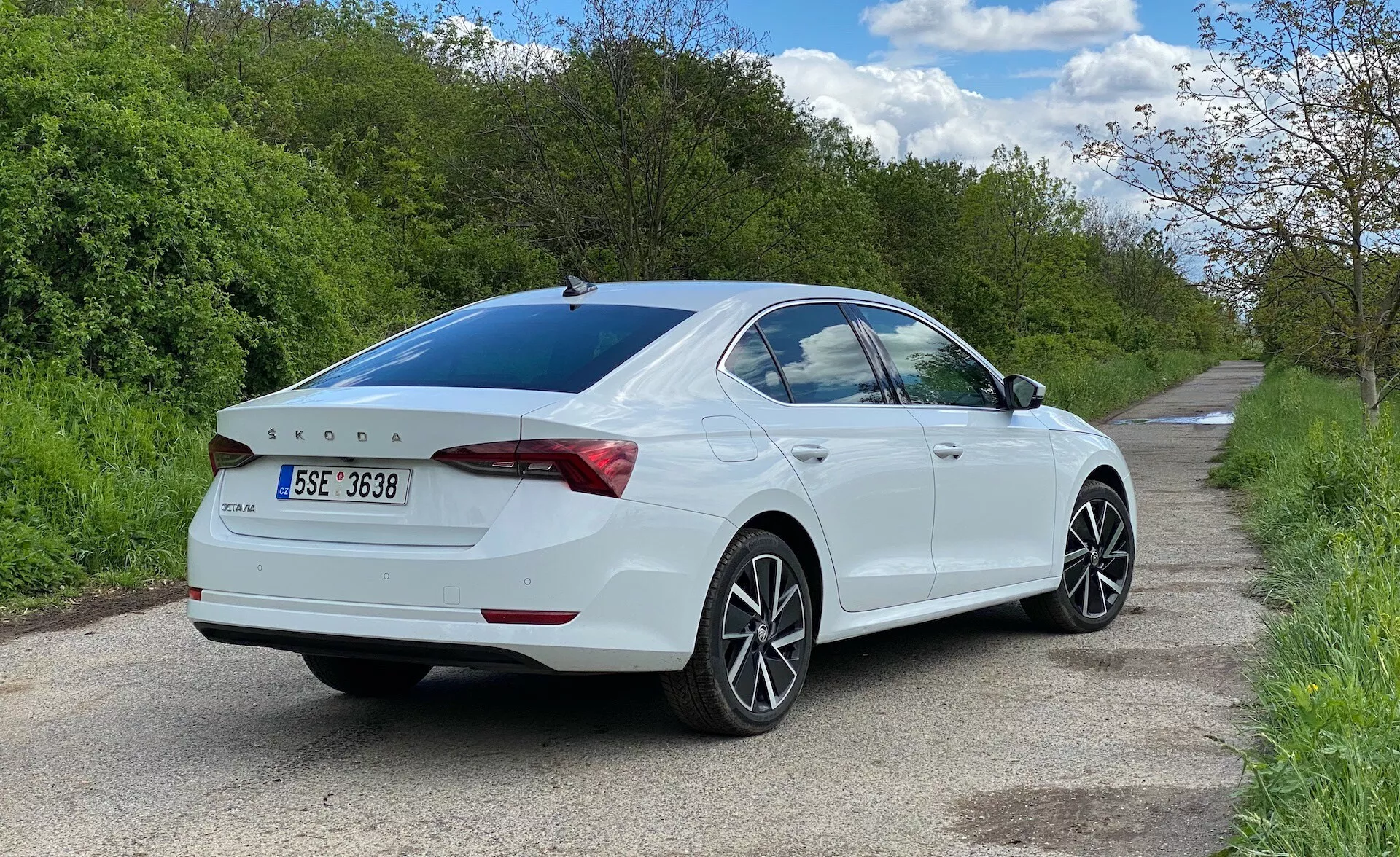 Vista traseira de três quartos de um sedan Skoda Octavia 2020 branco estacionado no lado direito de uma estrada estreita, mostrando o lado do passageiro a partir do nível do solo. O carro preenche a maior parte do quadro, destacando as linhas de design, as luzes traseiras envolventes até o porta-malas, spoiler integrado sutilmente, emblema traseiro, rodas de liga leve com múltiplos raios, vidro traseiro levemente escurecido e espelhos laterais com sinais de direção integrados.