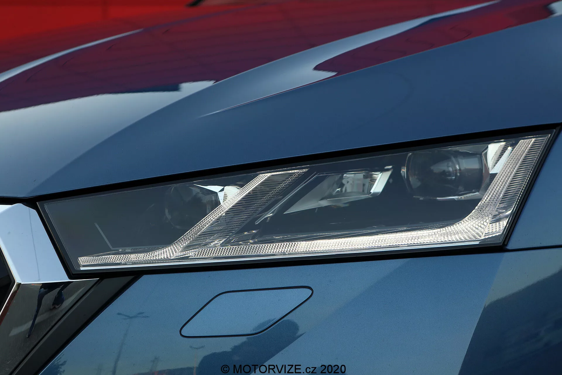 Uma foto de close-up do farol esquerdo no modelo limousine Škoda Octavia 2020, destacando seu design angular e a moderna tecnologia de iluminação LED ou laser. A fotografia, tirada de um ângulo baixo, mostra os refletores internos intrincados e os finos detalhes dentro do farol coberto por uma lente clara. A imagem de alta resolução também revela parte do capô e do para-lama com acabamento de pintura metálica, sugerindo um veículo premium ou bem mantido. Esta imagem enfatiza a iluminação automotiva avançada e o estilo agressivo associado a carros esportivos.