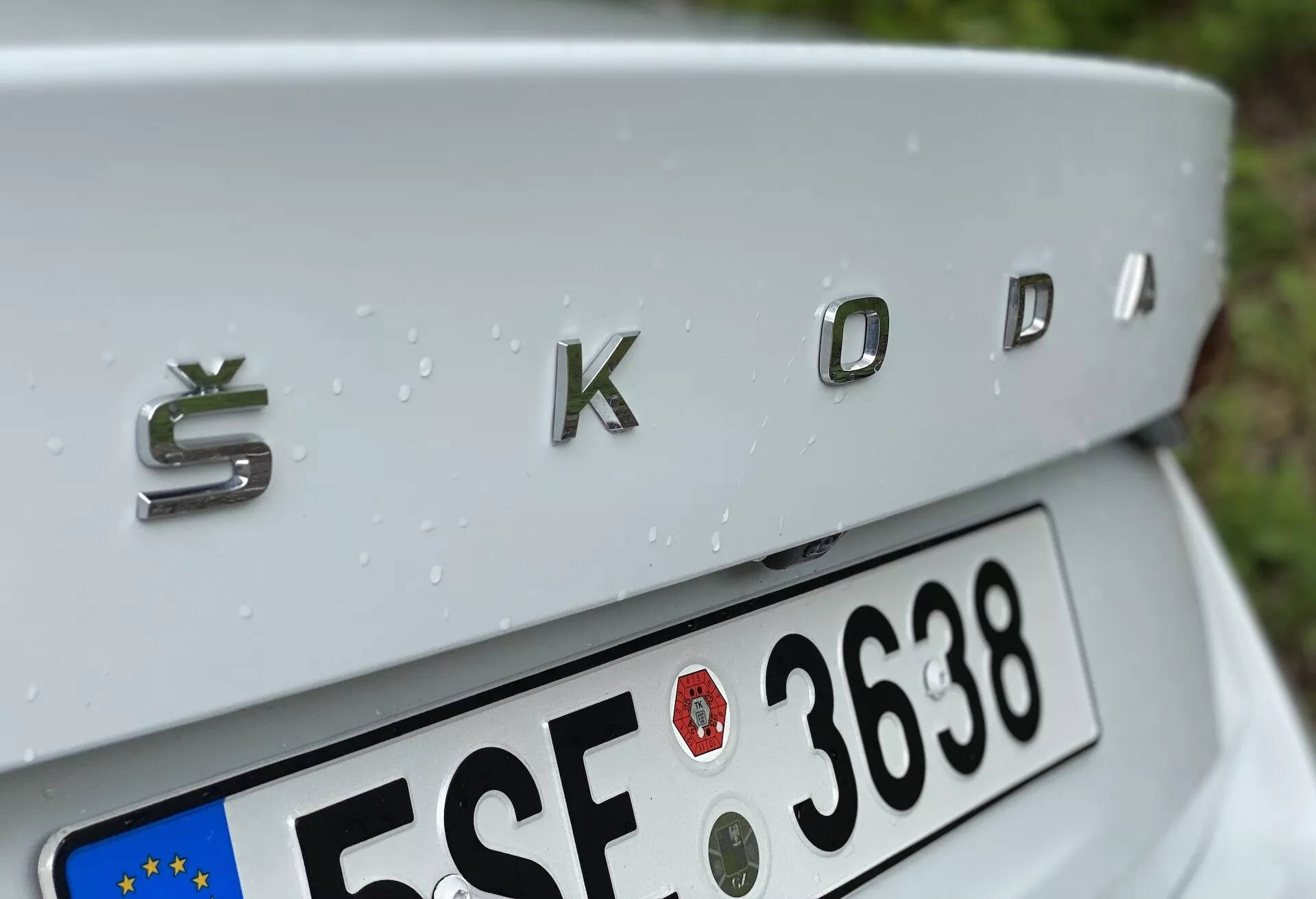 Close-up da seção traseira de um Škoda Octavia 2020 (octavia-limousine) focando no detalhe cromado 'ŠKODA' com acabamento metálico tridimensional. O ângulo da imagem ligeiramente de cima para baixo mostra a tampa do porta-malas suavemente inclinada, placa de licença estilo União Europeia parcialmente visível abaixo e gotas de chuva adornando a superfície prateada do carro, indicando chuva recente.