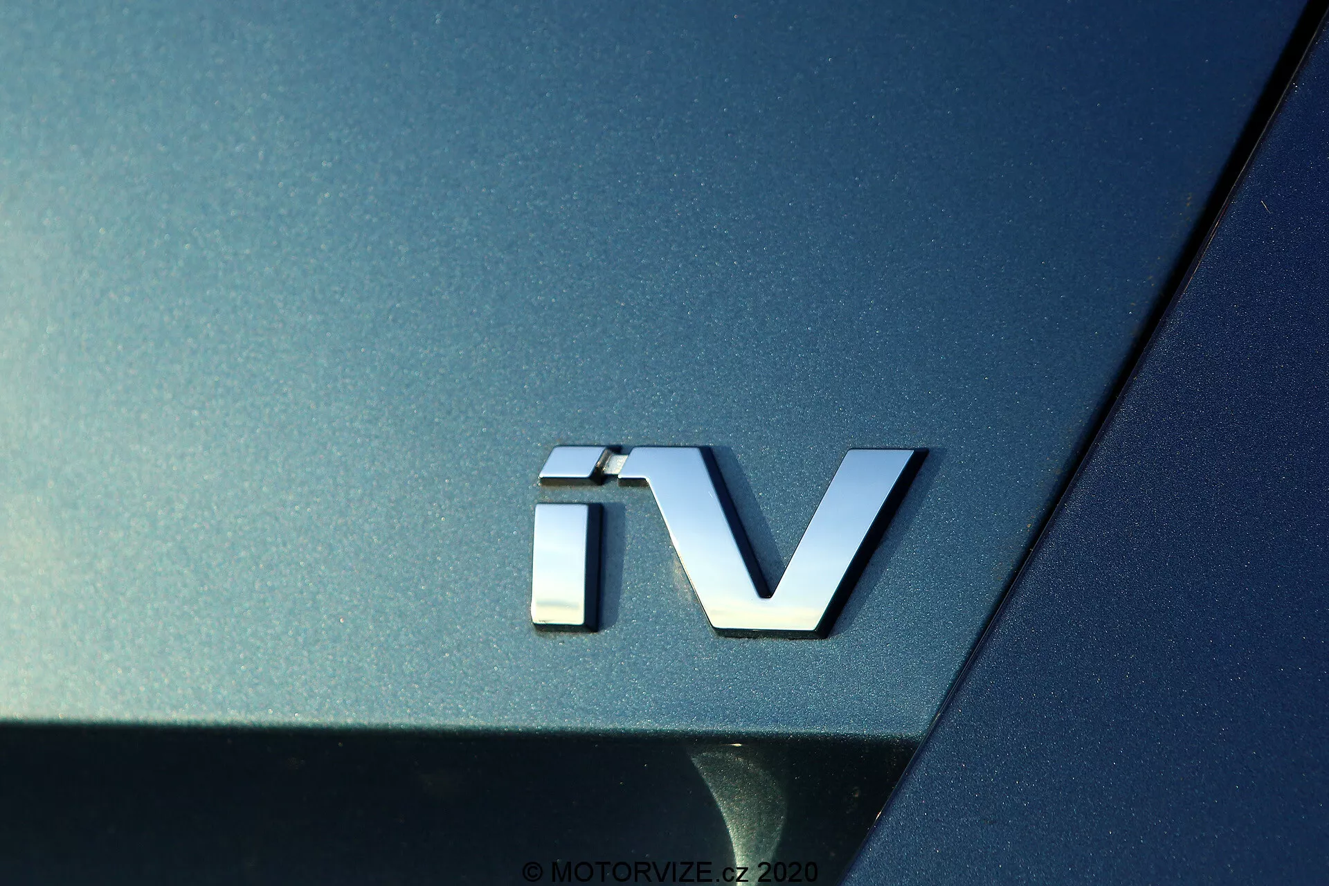 Close-up do emblema 'iV' do Skoda Octavia 2020, indicando um trem de força eletrificado, com um emblema 3D prateado contra a carroçaria metálica escura, destacando a superfície curva e as qualidades refletoras da seção traseira do carro.