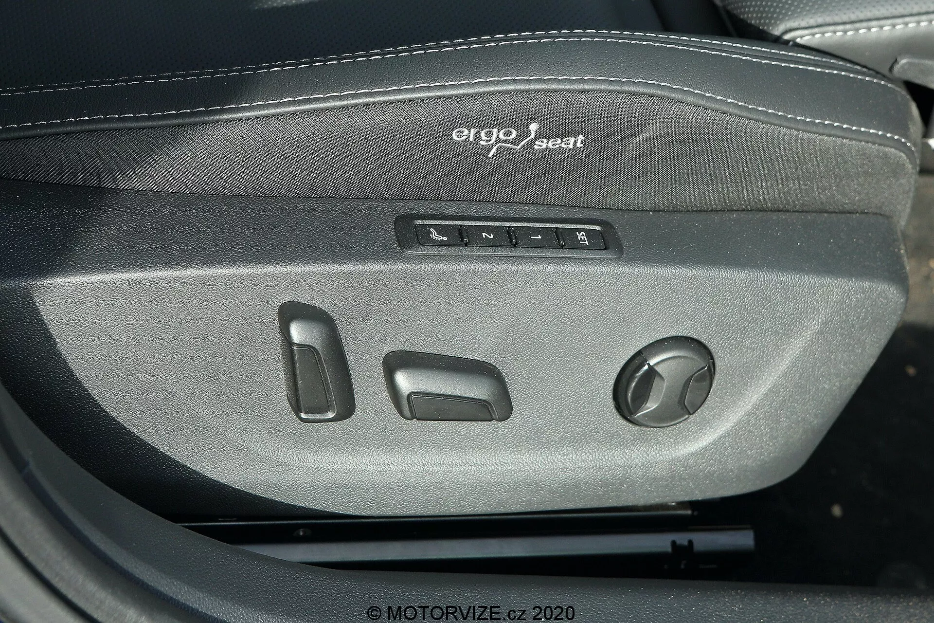Imagem de close-up mostrando os controles do sistema de assento ergo do Škoda Octavia Limousine 2020. A foto destaca vários botões e um botão rotativo para ajustar a posição, altura, inclinação e suporte lombar do assento do motorista ou passageiro. O texto 'ergo seat' está visivelmente bordado acima dos controles, enfatizando o design ergonômico. O assento é feito de tecido trançado cinza escuro, sugerindo um material durável e confortável adequado para veículos de médio a alto padrão.