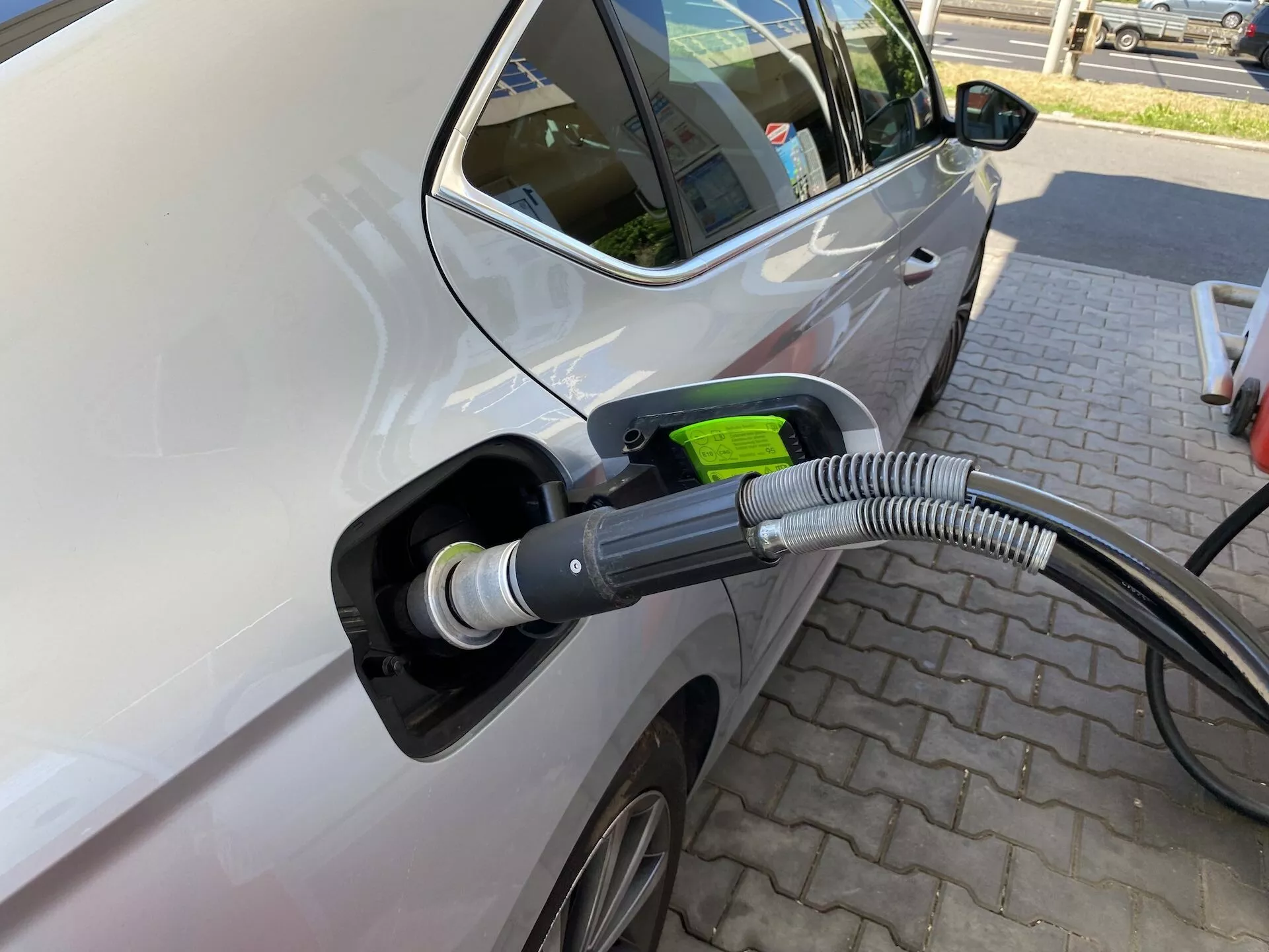Vista aproximada do lado esquerdo de um Skoda Octavia 2020 conectado a uma estação de carregamento elétrico, destacando a área do porto de carregamento com foco no cabo de carregamento e no porto. A imagem é tirada de um ângulo ligeiro da frente para a traseira, mostrando a porta do motorista da frente e parte da porta traseira, indicando que é um veículo de múltiplas portas. As características incluem um espelho lateral moderno, roda de liga parcialmente visível, cor clara do carro com acabamento brilhante, porto de carregamento aberto com capa protetora grossa e ondulada no cabo, e alça de plugue de cor verde simbolizando respeito ao meio ambiente. O carro está estacionado em uma área pavimentada com padrões de tijolos, sugerindo uma estação de carregamento dedicada, sem outros veículos próximos, enfatizando a ação de carregamento.