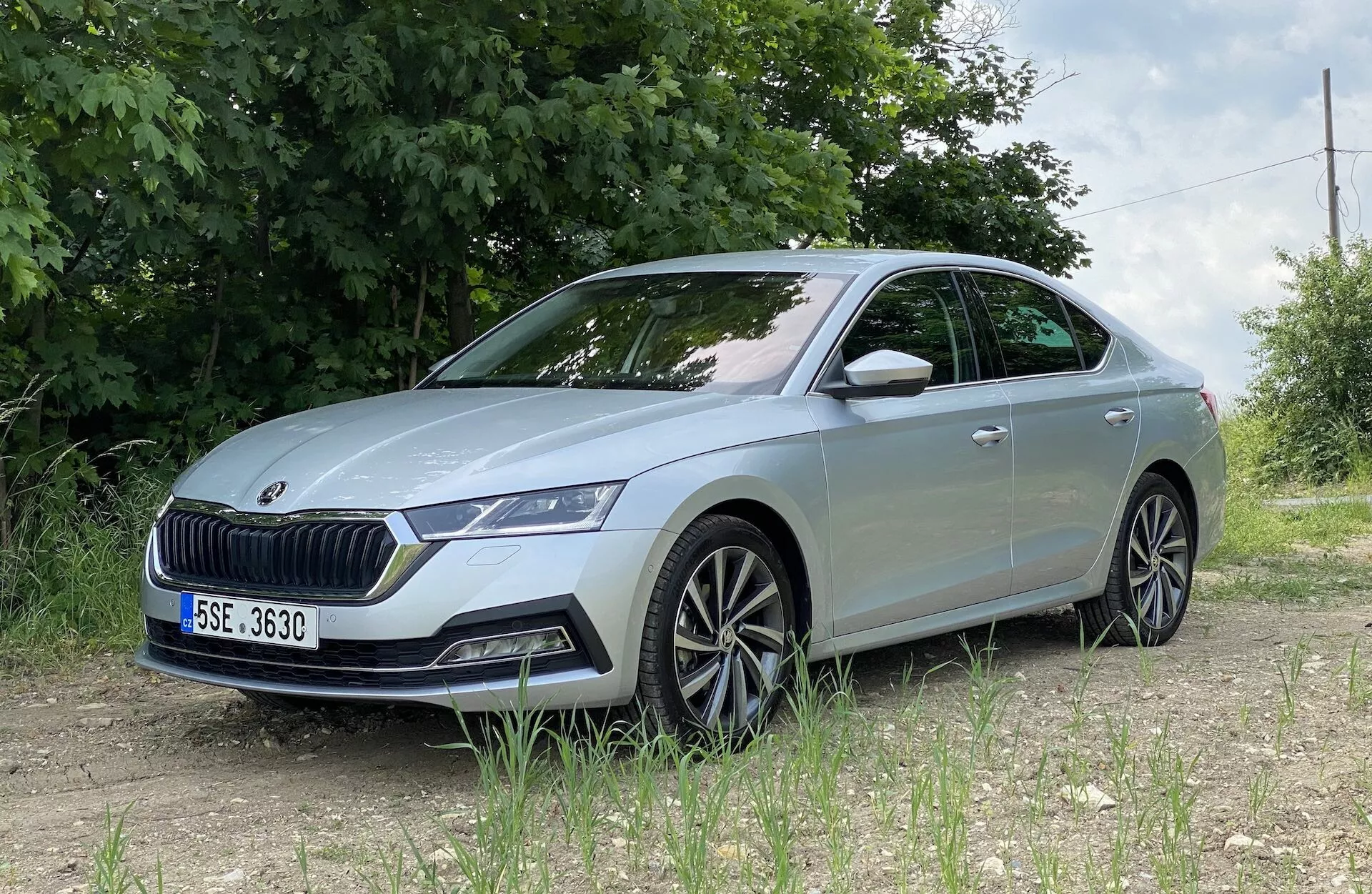 Skoda Octavia 2020 vista de um ângulo de três quartos frontal baixo, destacando o lado esquerdo dianteiro detalhado incluindo conjunto de faróis LED, grade proeminente, para-choque dianteiro com sensores, linhas esculpidas do capô, rodas de liga leve multi-raios, pisca integrado no retrovisor lateral e janelas ligeiramente escurecidas, posicionada em uma superfície não pavimentada com folhagem verde e cerca de arame em um cenário rural ou semi-rural.