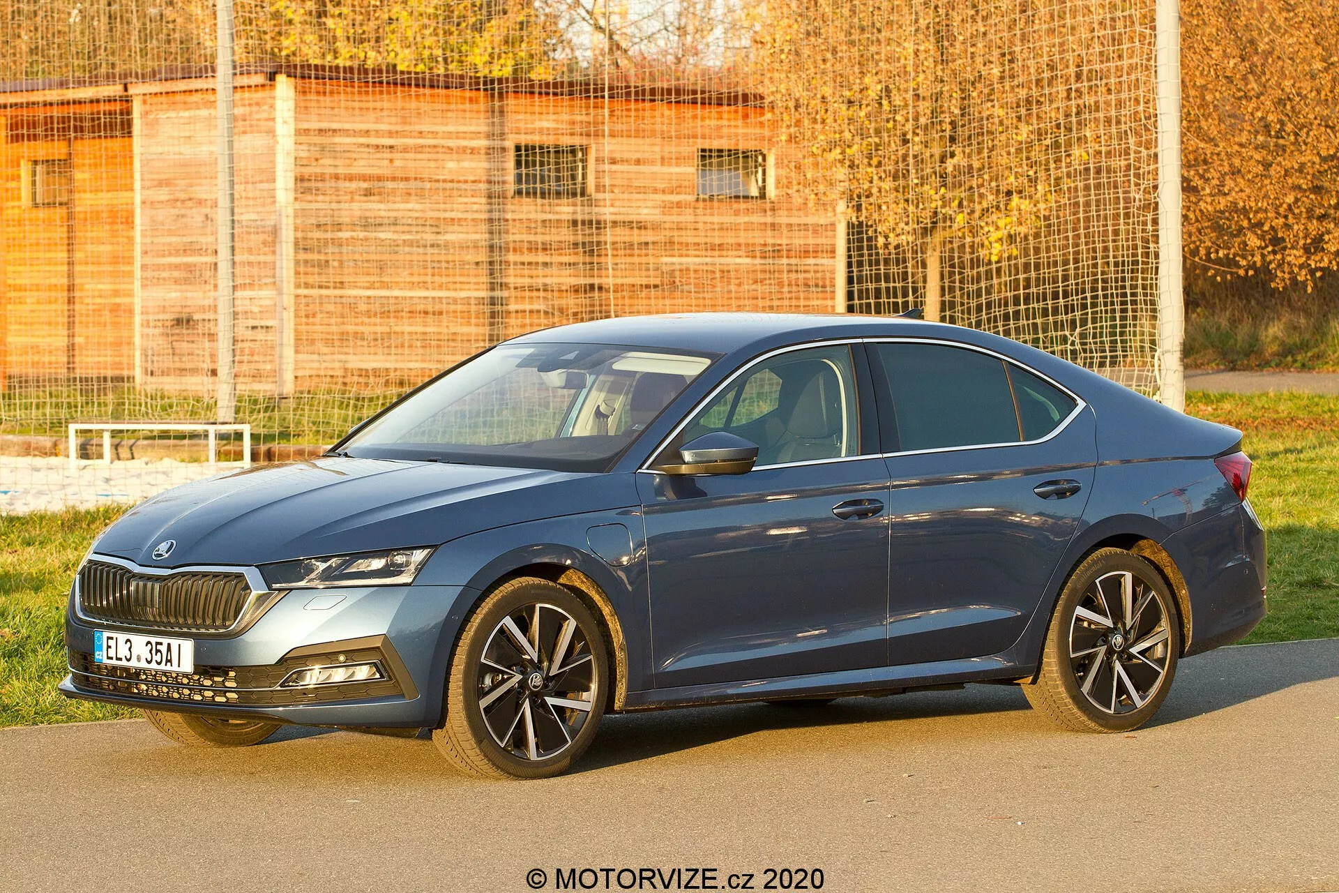 Imagem de um sedã Skoda Octavia 2020 de quatro portas, visto de um ângulo frontal de três quartos, mostrando sua grade frontal, faróis, capô, rodas, porta do motorista e uma parte da traseira. A foto captura o design distinto da grade, os faróis elegantes provavelmente equipados com sistemas de iluminação LED ou adaptativa, rodas de liga leve de múltiplos raios que sugerem esportividade ou luxo, pneus de perfil baixo e espelhos laterais com indicadores de direção integrados. As linhas aerodinâmicas da carroceria e a cor azul do carro são claramente visíveis, destacando elementos de design e tecnologia adequados para contextos automotivos e de veículos modernos.