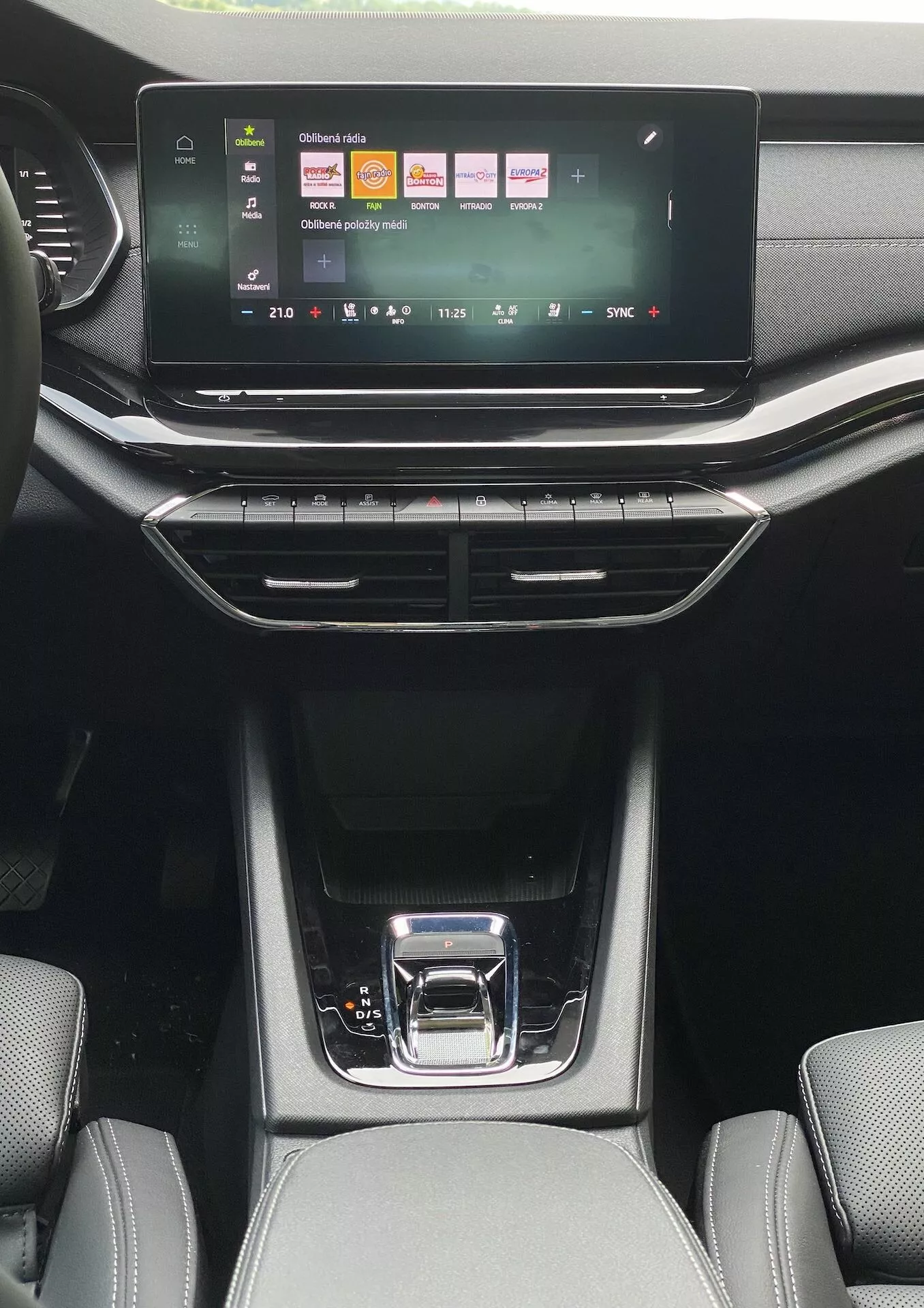 Perspectiva interior desde a área dos passageiros traseiros de um Skoda Octavia limousine 2020, mostrando o sistema de infotainment com tela sensível ao toque com ícones coloridos para mídia, telefone e configurações do veículo. Abaixo estão os botões de controle climático de zona dupla com um mostrador digital de temperatura, cercados por saídas de ar verticais e horizontais. O console central elegante apresenta uma alavanca de câmbio preta brilhante com uma alça prateada, compartimentos de armazenamento com tapetes de borracha e estofamento visível texturizado nos bancos dianteiros sob luz natural do dia.