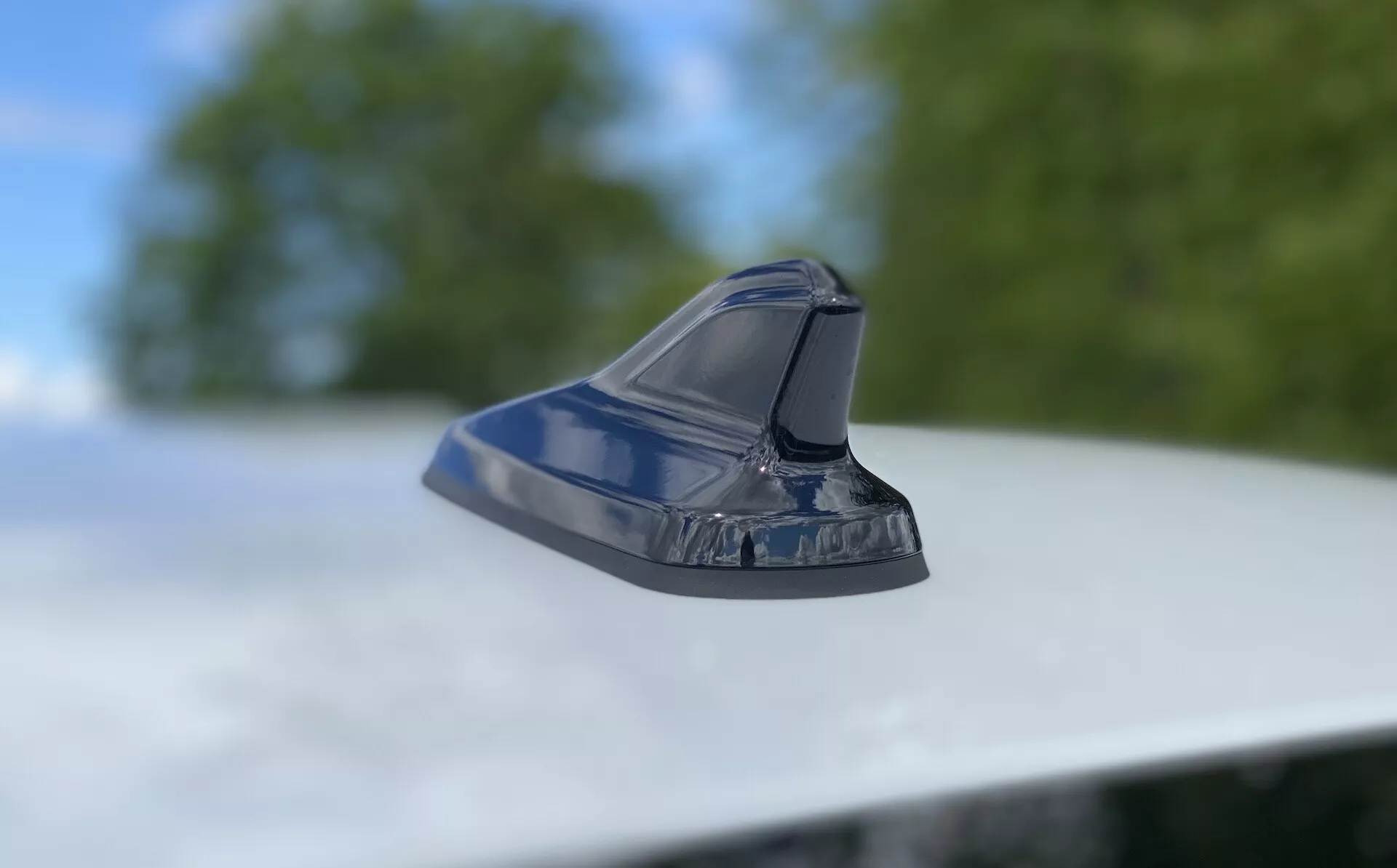 Close-up de uma antena estilo barbatana de tubarão no teto de um veículo, destacando o design automotivo moderno. Foto tirada de um ângulo baixo com a câmera ligeiramente inclinada para cima, focando na antena aerodinâmica e brilhante contra um fundo desfocado de folhagem e céu. O design elegante sugere funcionalidades como rádio FM/AM, navegação por satélite e tecnologia de comunicação avançada. O acabamento metálico reflete o exterior bem conservado do veículo.