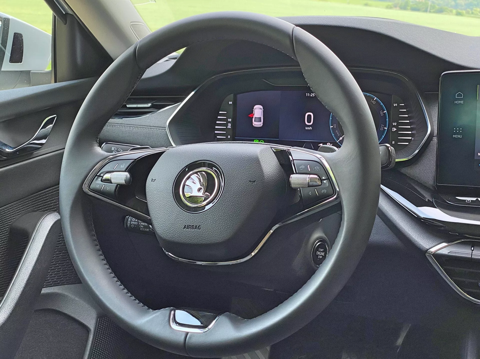 Interior do Skoda Octavia Limousine 2020 a partir da perspectiva do motorista, destacando o volante moderno de três raios revestido em couro com vários controles para volume, comandos de voz e configurações de controle de cruzeiro. O painel de instrumentos totalmente digital atrás do volante exibe um velocímetro estacionário e um indicador de portas abertas. O console central possui uma tela sensível ao toque para o sistema de infotainment para multimídia, navegação e configurações do veículo. O painel exibe um design moderno com materiais de alta qualidade.