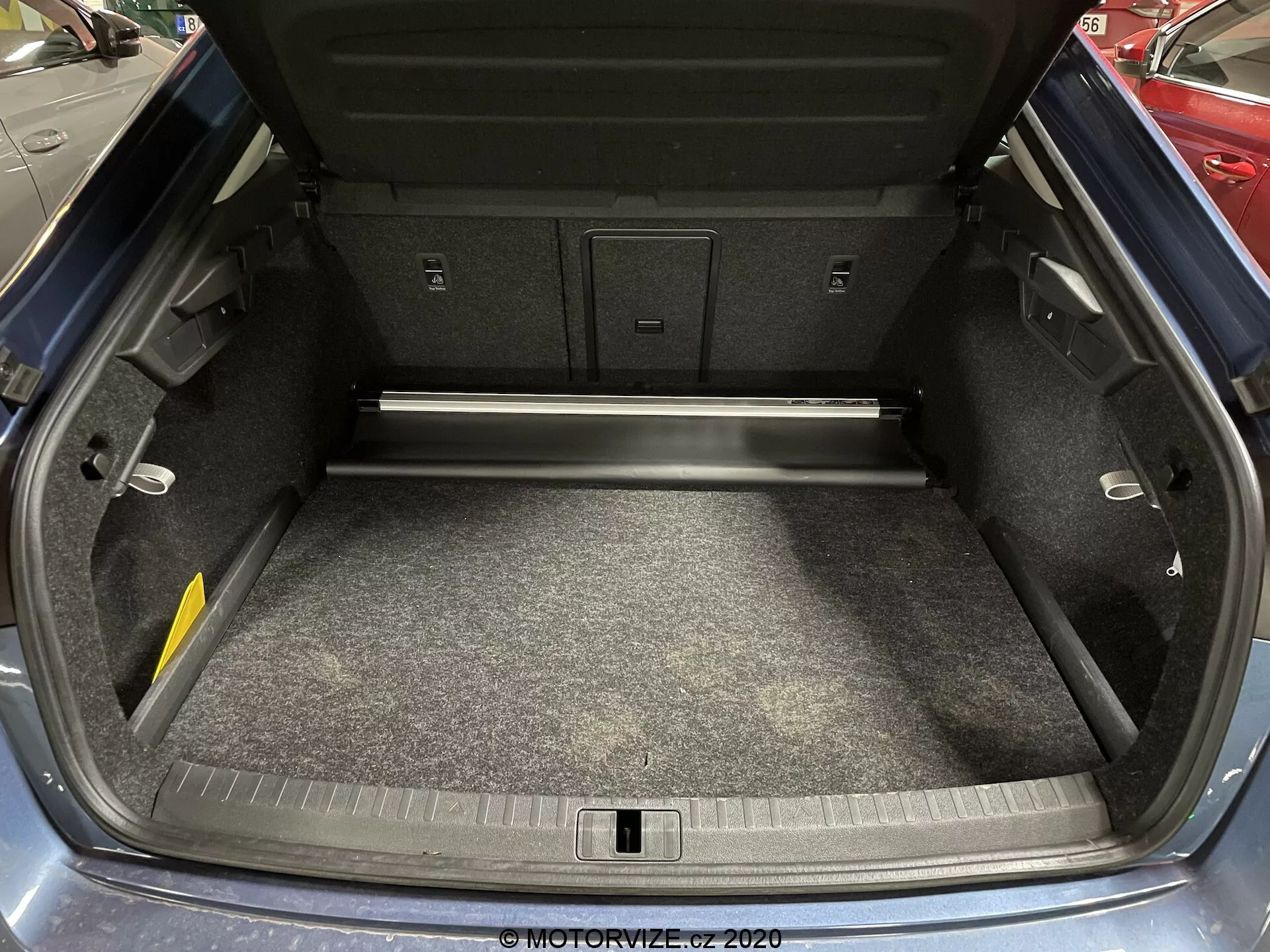 Imagem do porta-malas do Skoda Octavia 2020 com a tampa aberta, mostrando um interior espaçoso e limpo, piso acarpetado, revestido com painéis de material composto, uma passagem traseira para itens mais longos e gancho de carga visível. O parachoque traseiro apresenta marcas de desgaste menores.