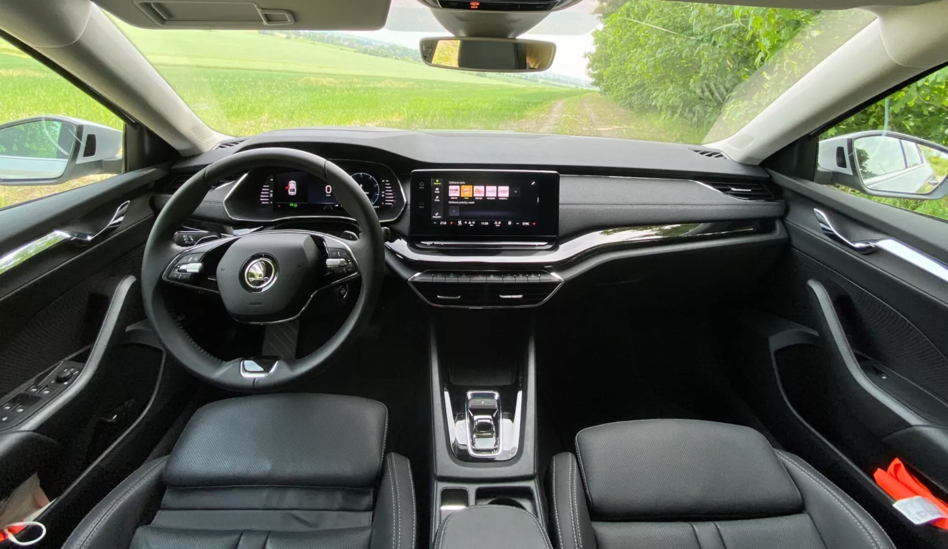 Vista interna de um Skoda Octavia 2020 a partir do banco traseiro mostrando o volante moderno com fundo plano e controles integrados, o painel de instrumentos digital, um grande sistema de infotainment com tela sensível ao toque em um painel elegante, poucos botões físicos, o carregador sem fio, bancos dianteiros ergonômicos de material semelhante ao couro com costura contrastante e um teto solar panorâmico permitindo a entrada de luz natural na cabine.