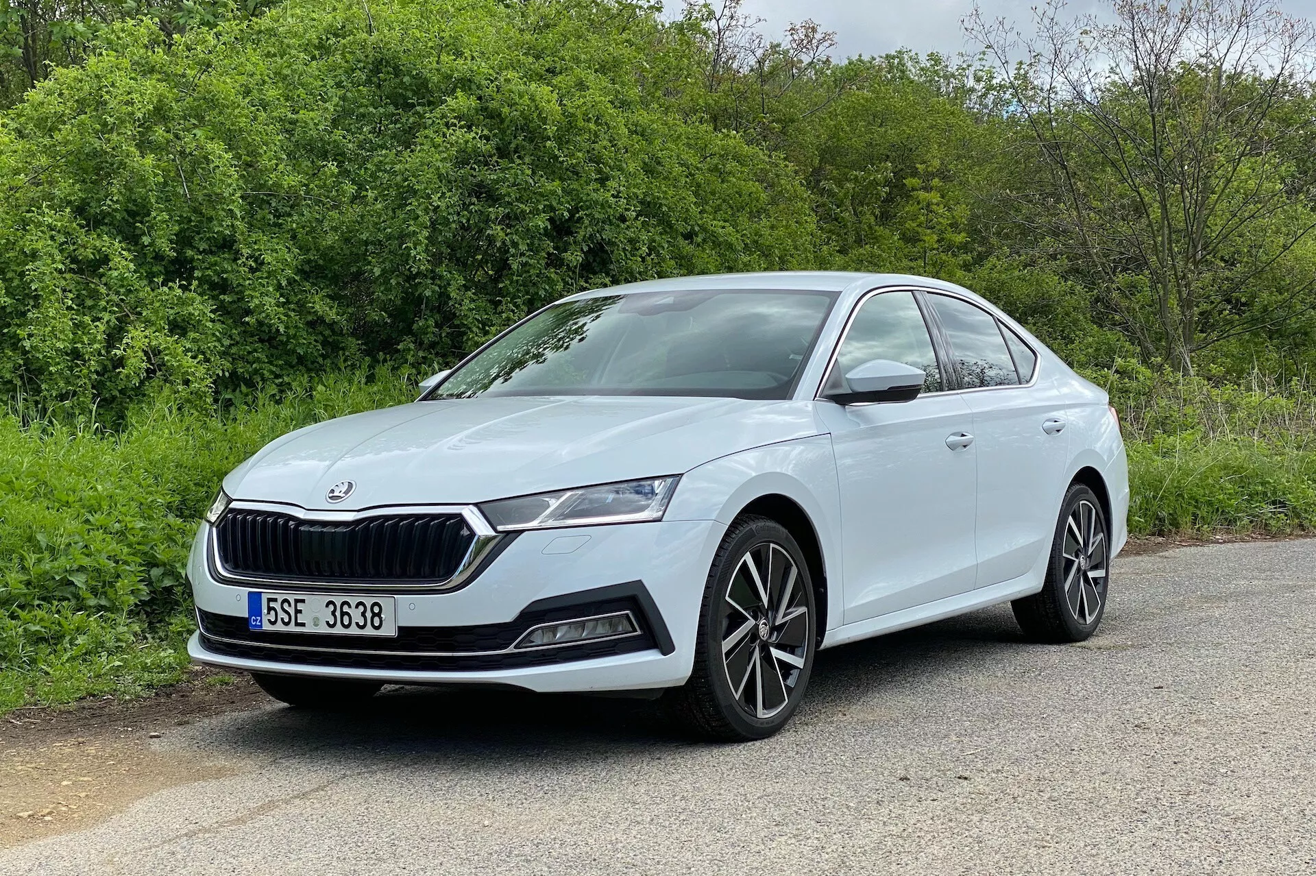 A imagem retrata um sedan Skoda Octavia 2020 em um ângulo de três quartos dianteiro, mostrando tanto a frente quanto o perfil lateral do lado do motorista. O carro, estacionado em uma superfície de asfalto com elementos naturais como árvores ou arbustos ao fundo, possui uma grade distintiva, faróis de LED elegantes, alojamentos de luzes de neblina e entradas de ar no para-choque. As rodas de liga leve multirraios e as linhas laterais suaves e aerodinâmicas melhoram sua aparência moderna e esportiva. A visão bem iluminada e desobstruída destaca as maçanetas da porta na cor da carroceria e a linha de caráter visível ao longo das portas, tornando-a uma excelente representação do design automotivo.