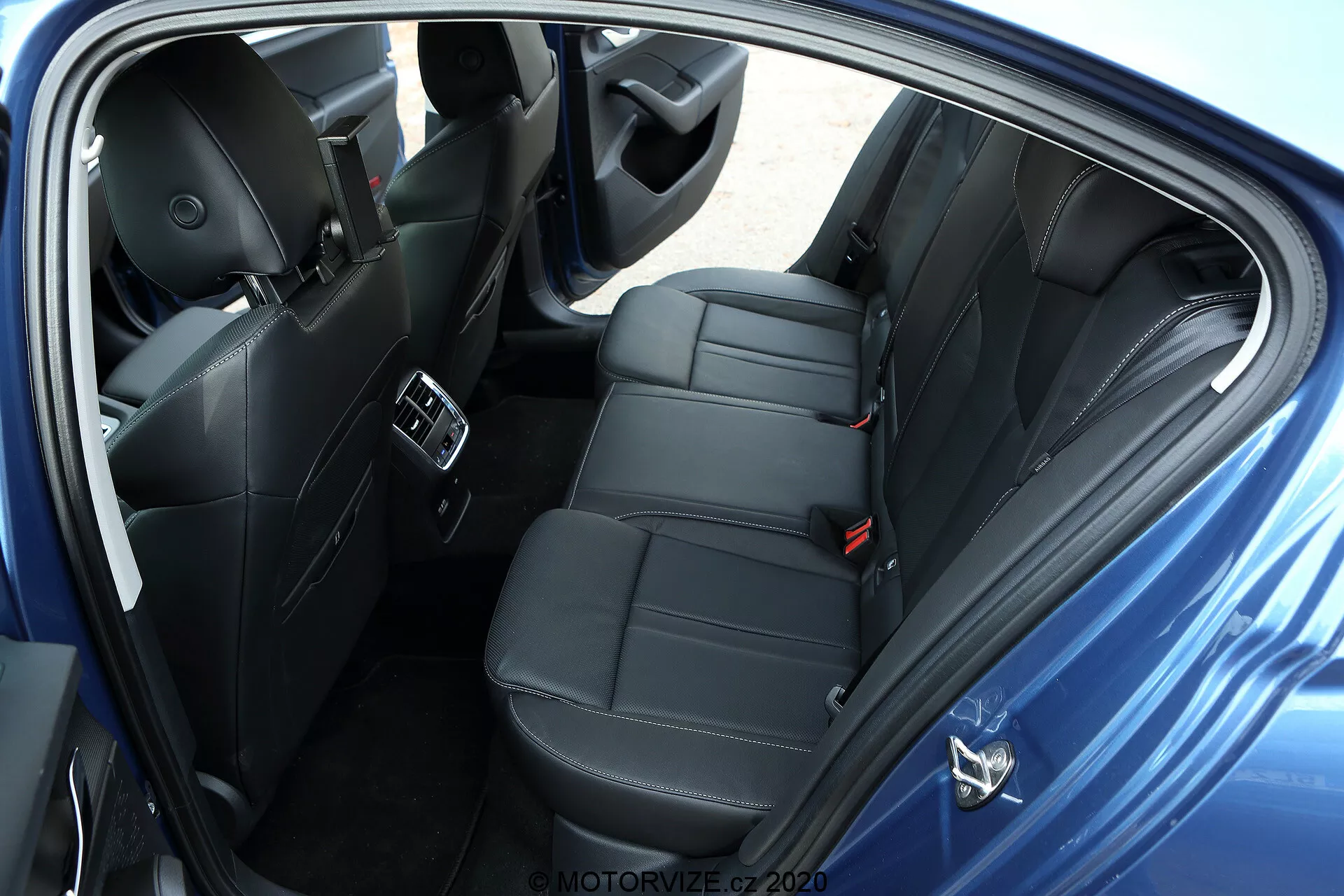 Vista aérea do compartimento traseiro para passageiros de um Skoda Octavia 2020 limousine, tirada do lado direito. A foto mostra uma porta aberta com assentos modernos de couro ou material semelhante a couro preto com costura de luxo. O banco traseiro é projetado para acomodar confortavelmente dois passageiros com apoios de cabeça individuais, indicando um modelo mais esportivo ou focado no conforto. A imagem destaca as aberturas de controle de clima no console central, portas USB para carregamento de dispositivos, alto-falantes do sistema de áudio avançado no painel da porta, controles de vidros elétricos, cintos de segurança visíveis, incluindo um cinto de três pontos para o assento central, e tapetes carpetados. O esquema de cores escuras do interior aponta para um veículo premium bem equipado sem itens pessoais visíveis, em posição estacionada com fundo neutro.