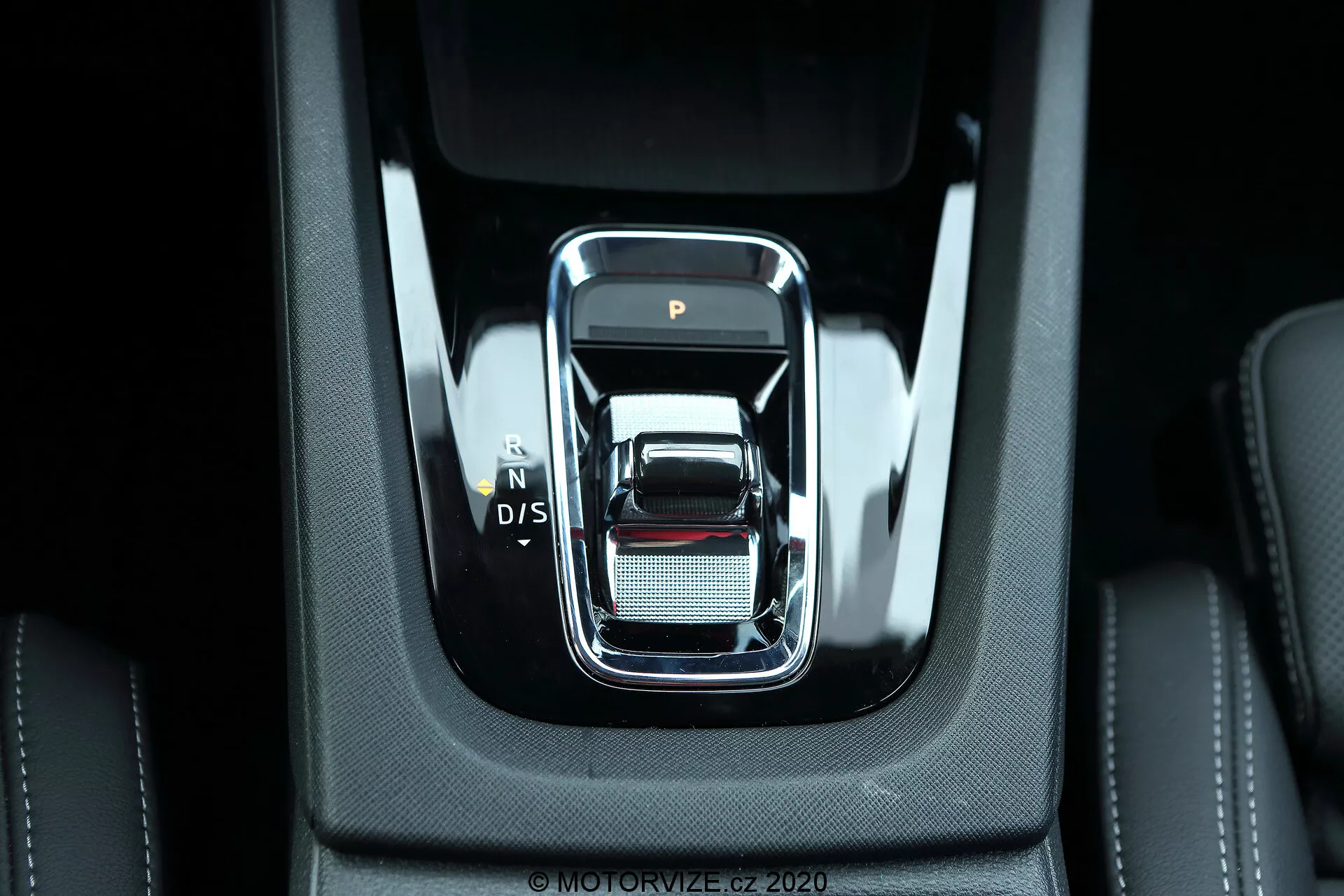 Vista de perto do moderno seletor de marchas no console central de um Skoda Octavia Limousine 2020. O seletor de marchas tem um design elegante, retangular com bordas arredondadas e contorno cromado, apresentando as posições 'P', 'N', 'D/S'. Cercado por materiais pretos de alta qualidade com costuras visíveis e uma superfície prateada com uma textura de malha fina, sugerindo luxo e detalhamento. A iluminação brilhante ilumina o interior robusto e coeso, indicativo de recursos eletrônicos avançados e tecnologias automotivas modernas.