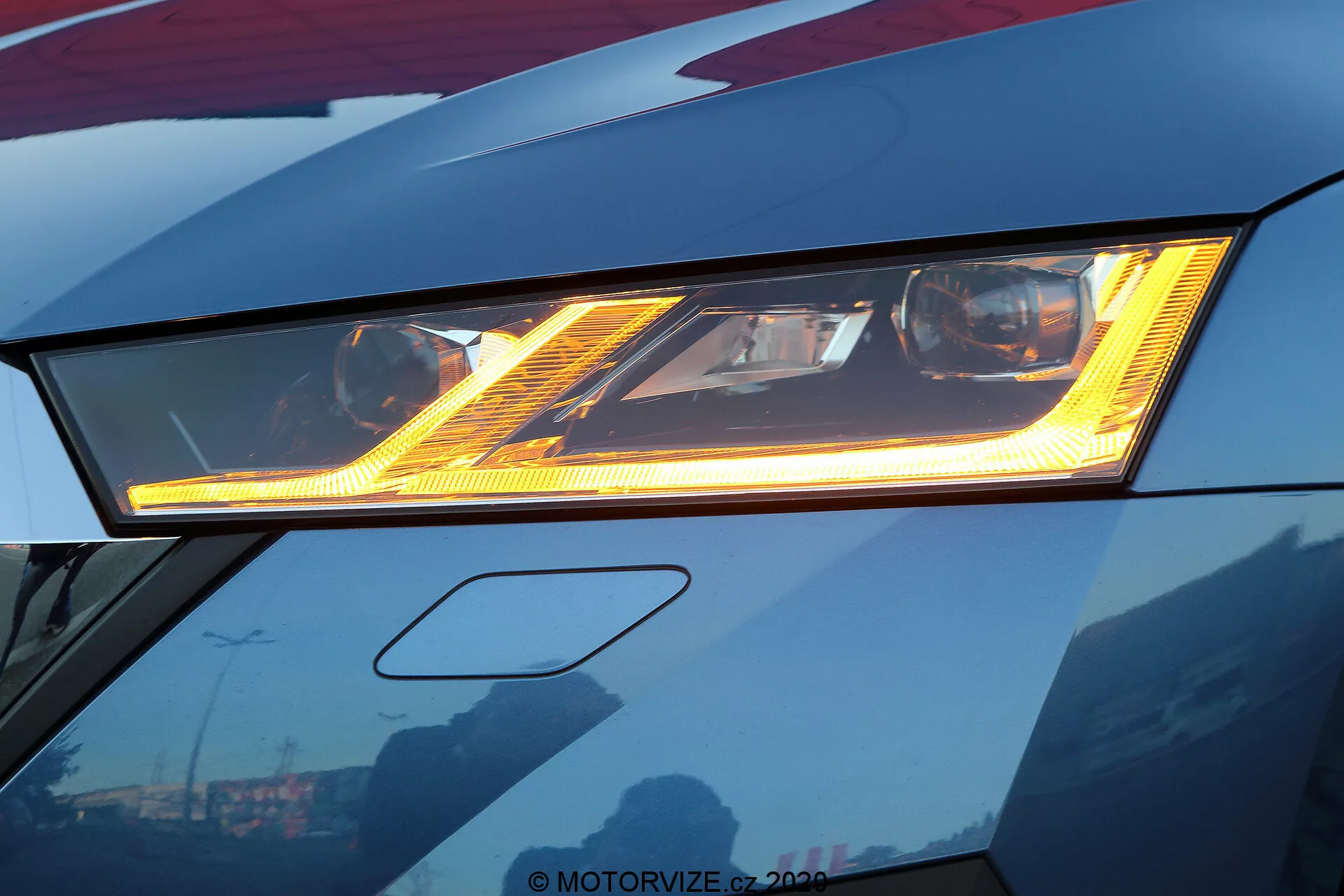 Close-up do farol LED avançado de um Skoda Octavia 2020 vermelho escuro, destacando sua iluminação brilhante e padrões de luz distintos. O design sofisticado apresenta uma luz diurna (DRL) sob o farol principal, com a carroceria elegante exibindo bordas afiadas. Reflexos do céu e dos prédios na superfície bem polida sugerem um acabamento premium. A moldura revela ligeiramente o para-choque dianteiro, focando na inovação do design automotivo.