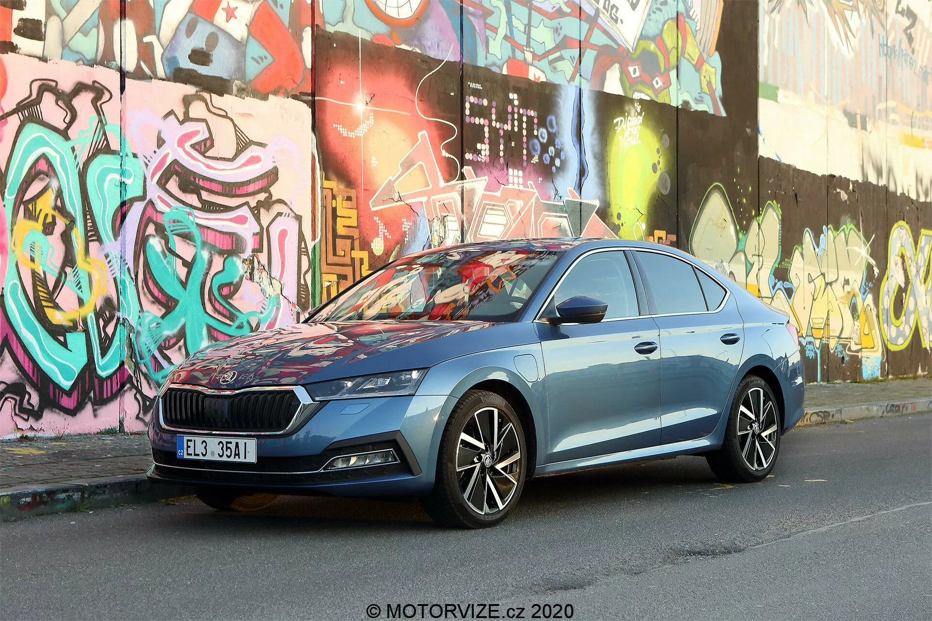 A imagem exibe um Skoda Octavia Limousine 2020 estacionado em uma rua urbana com paredes cobertas de graffiti ao fundo. Capturada de um ângulo frontal três-quartos ligeiramente baixo, destaca a grade substancial do carro com um padrão vertical e emblema central, faróis LED elegantes, rodas de liga leve multi-raios, linhas de corpo dinâmicas, espelhos laterais com indicadores de direção integrados, moldura cromada das janelas e pintura metálica azul. O ambiente urbano realça o design moderno e a avançada tecnologia automotiva do veículo.