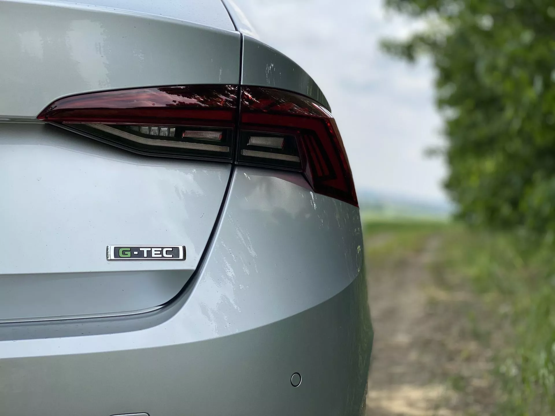 Vista detalhada e em close da seção traseira direita de um Skoda Octavia 2020 limousine. A foto captura os elementos de design da tampa do porta-malas, a lanterna traseira de LED e parte do para-choque traseiro de um ângulo oblíquo e ligeiramente elevado. O emblema 'G-TEC' na tampa do porta-malas sugere uma tecnologia ou nível de acabamento específico. A pintura metálica destaca o exterior polido do carro. O fundo desfocado proporciona um cenário rural que enfatiza as características e o design do veículo.