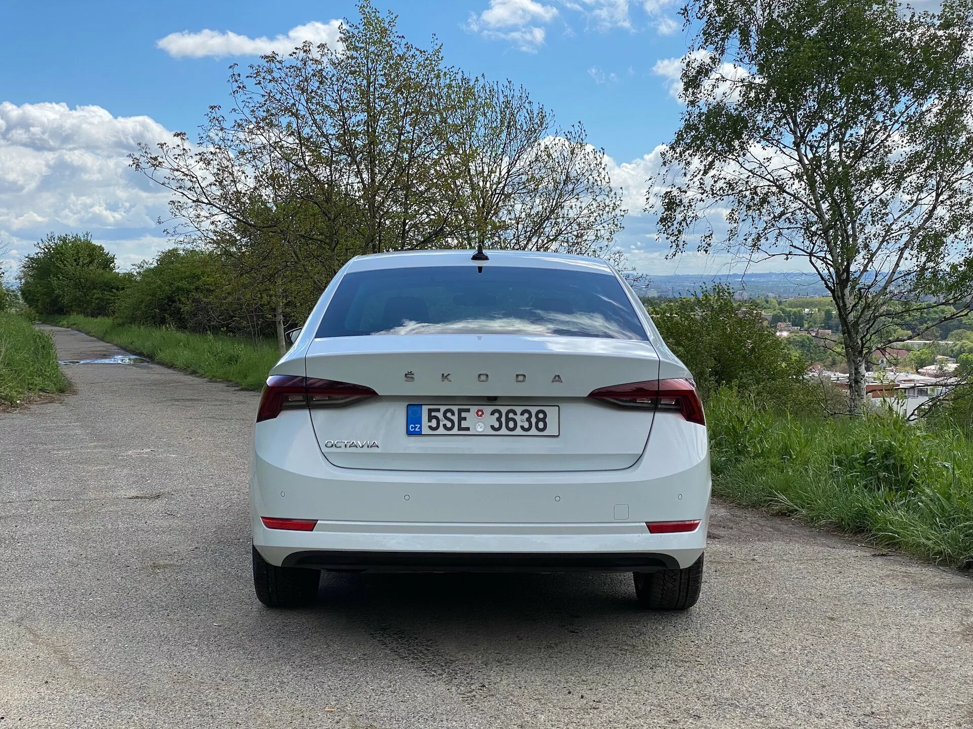 Vista traseira de um modelo Škoda Octavia 2020 limousine branco, estacionado em uma estrada estreita de asfalto em um ambiente rural. A imagem, tirada do nível do solo em ângulo reto direto, mostra a parte traseira do carro, incluindo a tampa do porta-malas, o para-choque traseiro, as luzes traseiras, a placa de matrícula, o para-brisa traseiro e a marca Škoda exibida proeminentemente no centro da tampa do porta-malas com 'Octavia' à esquerda. Ao redor do veículo há árvores e um céu azul nublado, com uma cidade ou paisagem distante visível abaixo do horizonte.