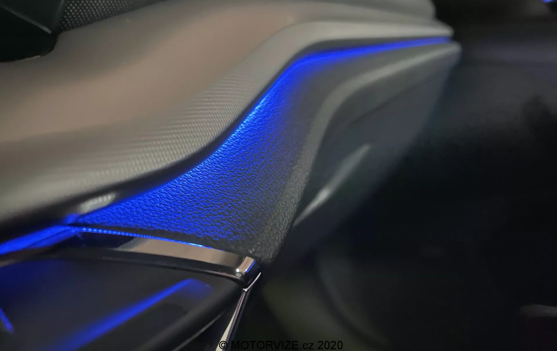 Detalhe interior em close-up do Skoda Octavia Limousine 2020, destacando a faixa de iluminação ambiente azul ao longo da porta ou painel do carro. O design elegante com curvas suaves se integra perfeitamente com a superfície de couro, enfatizando a sensação de luxo sob a iluminação suave. Tirada em um ambiente escuro, a fotografia acentua a iluminação azul e as texturas dos materiais, mostrando a tecnologia de iluminação ambiente frequentemente controlada através do sistema de infoentretenimento do veículo.