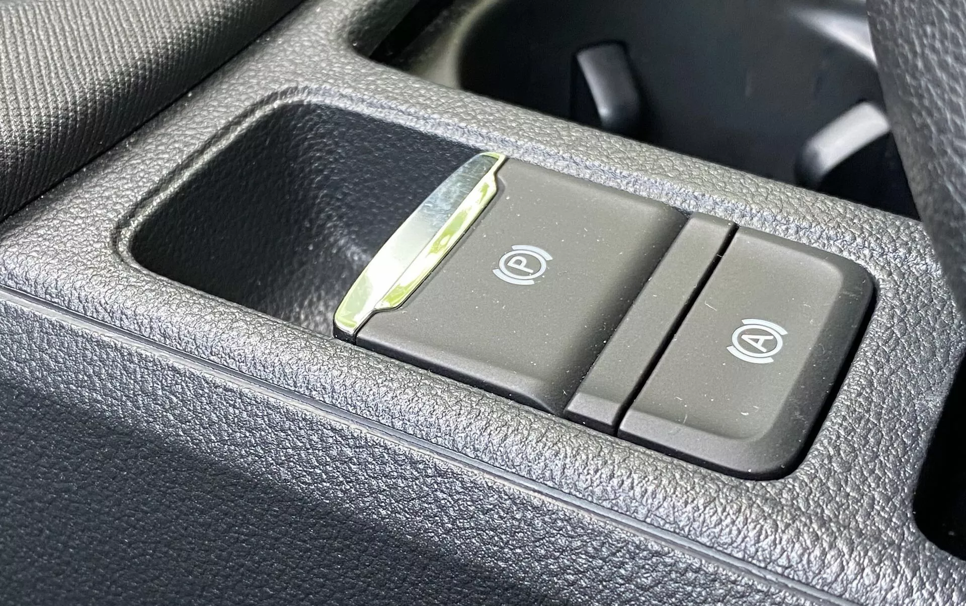Close-up do painel de controle do Skoda Octavia 2020 dentro do veículo, mostrando botões dos assentos aquecidos e ventilados em um material escuro, granulado e semelhante ao couro. O botão esquerdo apresenta um ícone de assento aquecido e o botão direito exibe um ícone de assento ventilado, com um pequeno indicador LED entre os botões.