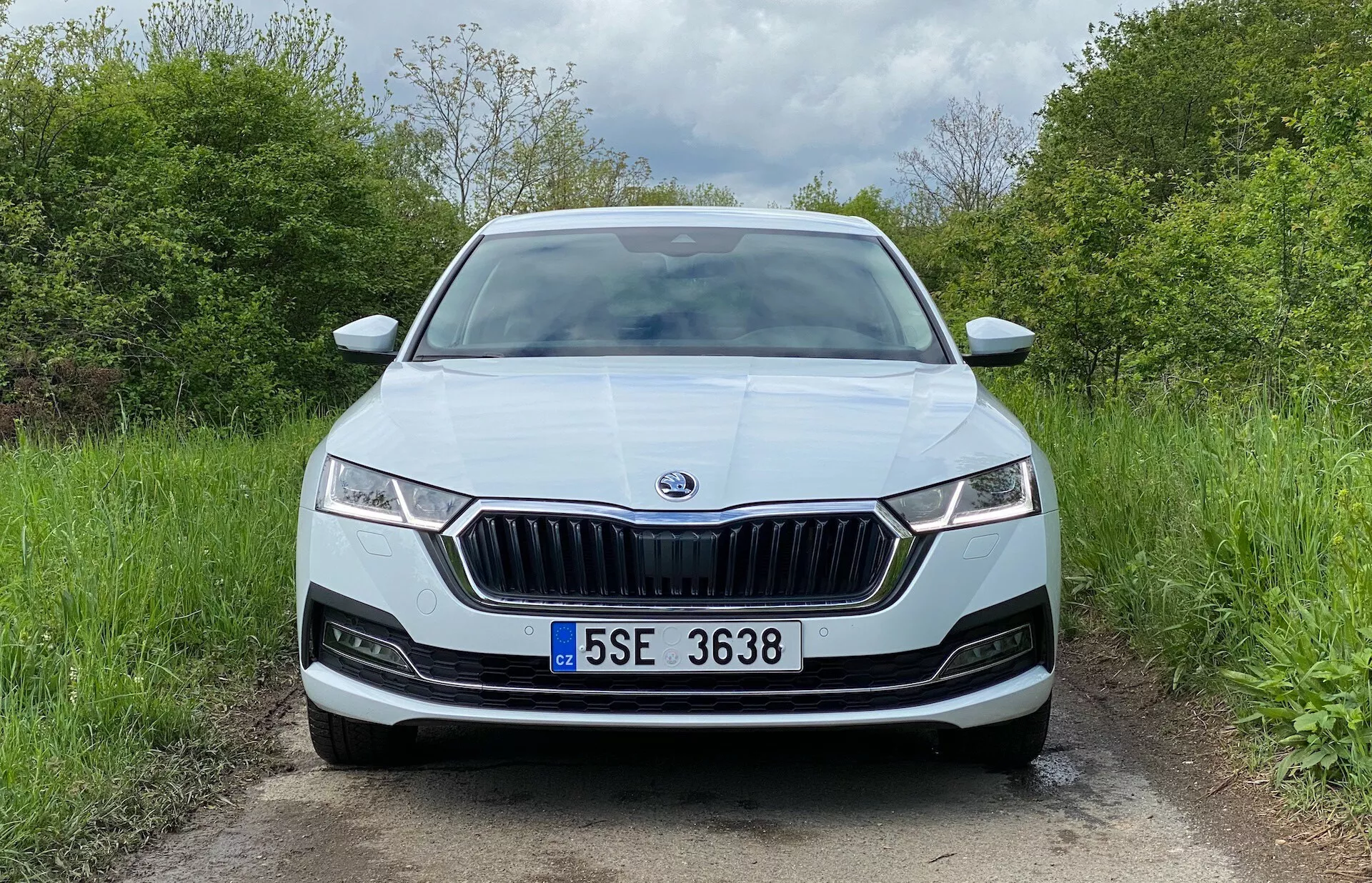 Vista frontal de um sedã Skoda Octavia 2020 em uma estrada rural destacando a grade frontal proeminente com lâminas verticais e contorno cromado, faróis LED estreitos com luzes diurnas, emblema da marca centralizado, sensores de estacionamento potenciais no para-choque, placa centralizada, acabamento de pintura metálica, entradas de ar e possivelmente luzes de nevoeiro.