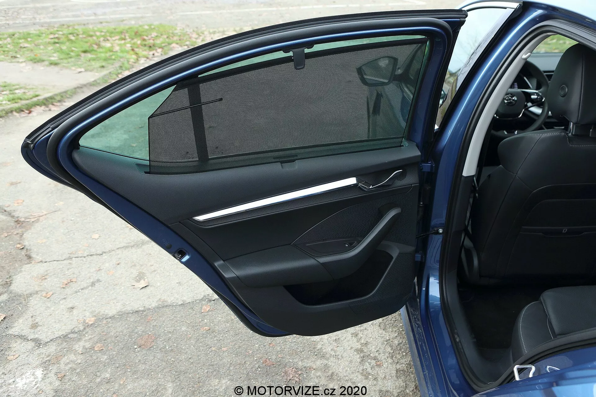 Vista aproximada da porta dianteira do passageiro aberta do Skoda Octavia 2020 limusine, destacando o painel interior da porta de cor escura com puxador metálico, painel de controle para vidros elétricos, compartimento de armazenamento, grade de alto-falante, soleira parcialmente visível e janela com cortina de sol.