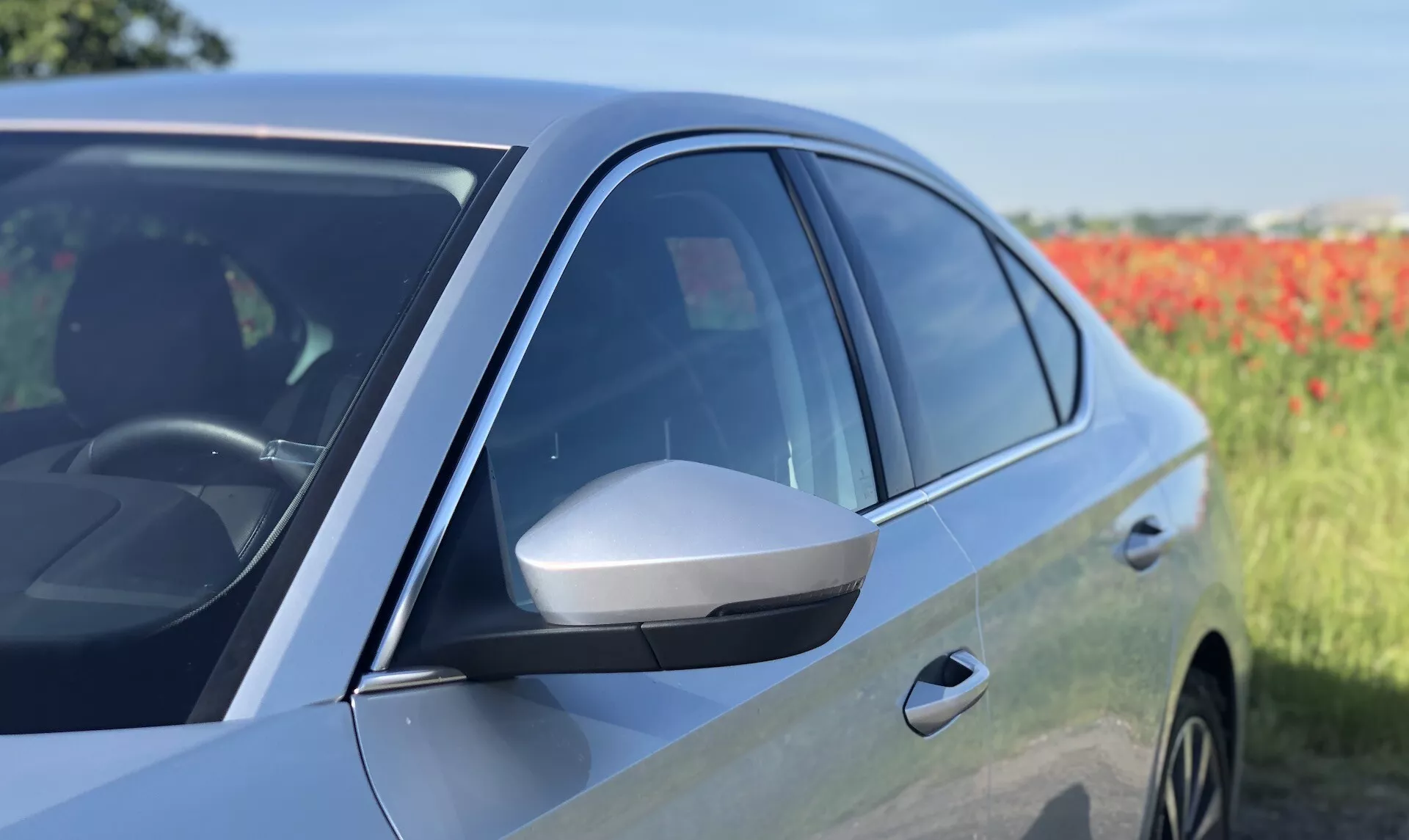 Vista lateral aproximada de um sedã moderno Škoda Octavia 2020, mostrando a porta frontal do lado do motorista e o espelho lateral. A imagem enfatiza o espelho lateral aerodinâmico com luz de seta integrada, janelas escurecidas, acabamento de pintura brilhante e design aerodinâmico do meio para a frente. O reflexo no painel da porta e parte do arco da roda dianteira sugerem a presença de rodas de liga leve. A foto destaca características de design avançadas e potenciais tecnologias do veículo contra um fundo natural ligeiramente desfocado.