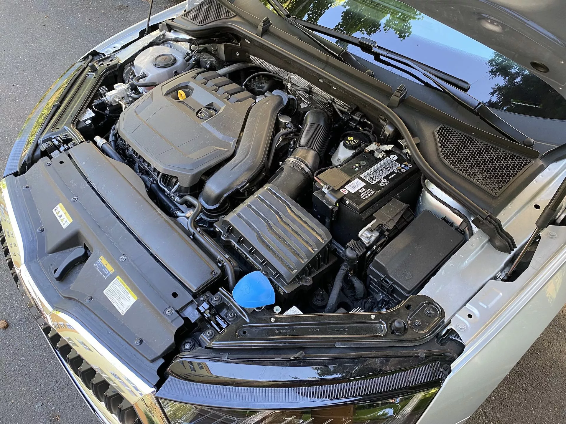 Vista aproximada do compartimento do motor do Skoda Octavia 2020 limousine com capô aberto, mostrando vários componentes, incluindo a tampa do motor centralmente enquadrada, a bateria à direita, um componente retangular com aletas de resfriamento à esquerda e um reservatório de fluido com tampa azul. A imagem, capturada de um ângulo descendente, ligeiramente deslocado, também mostra a carenagem de plástico preto na base do para-brisa, destacando a complexidade técnica do compartimento do motor automotivo.