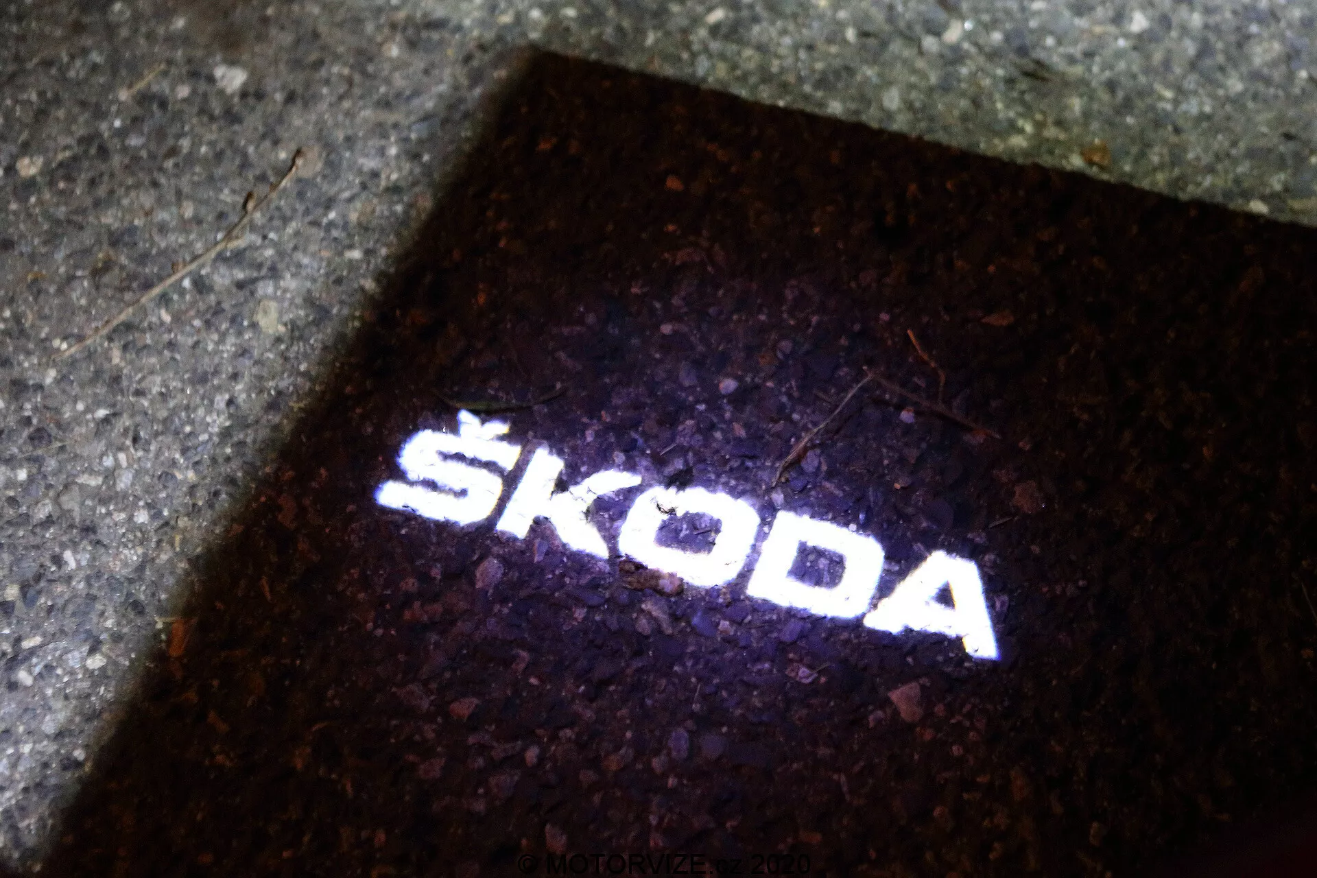 Projeção do logotipo Škoda no chão. A fonte de luz, provavelmente localizada no exterior do Škoda Octavia 2020 (octavia-limousine), cria um 'puddle light' que ilumina o chão quando a porta do veículo é aberta. Esta projeção de luz branca nítida e clara, em fundo escuro, serve para fins funcionais e de marca, indicando a presença de tecnologia LED moderna nos acessórios do veículo.