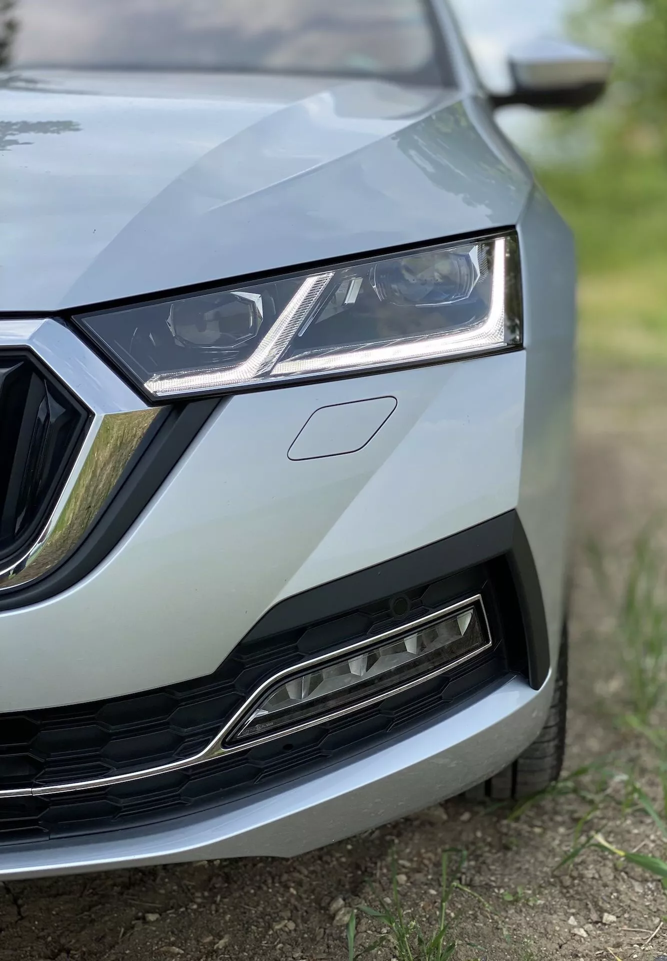 Close-up da seção frontal esquerda do Skoda Octavia 2020 (Octavia Limousine) mostrando detalhes do centro da grade ao farol esquerdo, parte do capô e uma porção do para-choque dianteiro e para-lama. A imagem, tirada de um ângulo baixo, destaca o design elegante dos faróis com luzes diurnas em LED, a grade com acentos cromados e a grade inferior com seu acabamento metálico moderno. O foco está nos detalhes do carro com uma profundidade de campo rasa, criando uma aparência profissional e de alta qualidade.