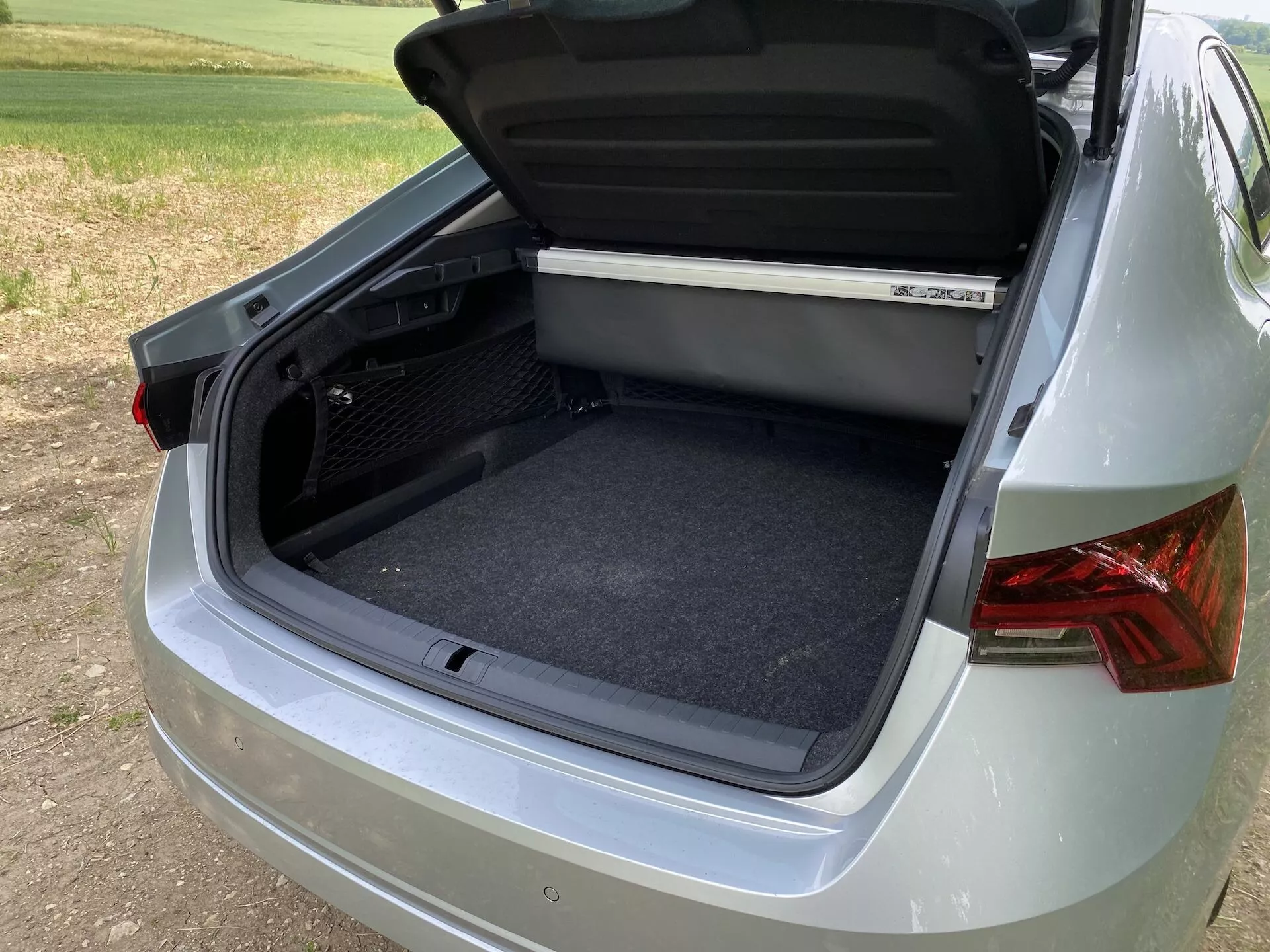 Close-up em três quartos do compartimento de bagagem traseiro de um Škoda Octavia 2020, focando no espaço de armazenamento aberto com um interior limpo e revestido de carpete. O ângulo captura parte do lado direito e traseiro do carro, mostrando luzes traseiras de design moderno, pintura metálica lisa e um para-choque parcialmente visível. A parte inferior da tampa do porta-malas inclui uma alça ergonômica e uma prateleira é vista no topo do espaço de carga. O ambiente sugere um cenário ao ar livre com iluminação suave durante o dia.