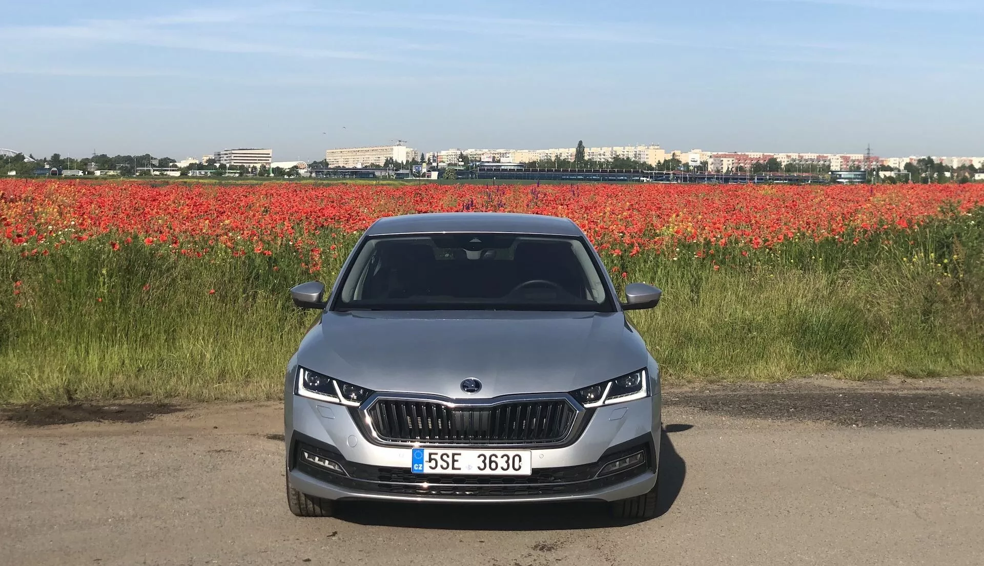 Vista frontal de um Skoda Octavia Limousine 2020 em uma estrada ou trilho de terra, mostrando sua fachada frontal com uma grade distinta em forma de rim ladeada por faróis elegantes de LED ou adaptativos. O para-choque dianteiro aerodinâmico apresenta recortes para luzes de neblina e entradas de ar. O capô tem linhas de contorno que sugerem um design estético e aerodinâmico. O para-brisa totalmente visível pode sugerir recursos tecnológicos avançados, como displays head-up ou sistemas de assistência ao motorista. Um carro de cor clara com placa de licença montada centralmente e sensores de estacionamento, contra um fundo de flores vermelhas.