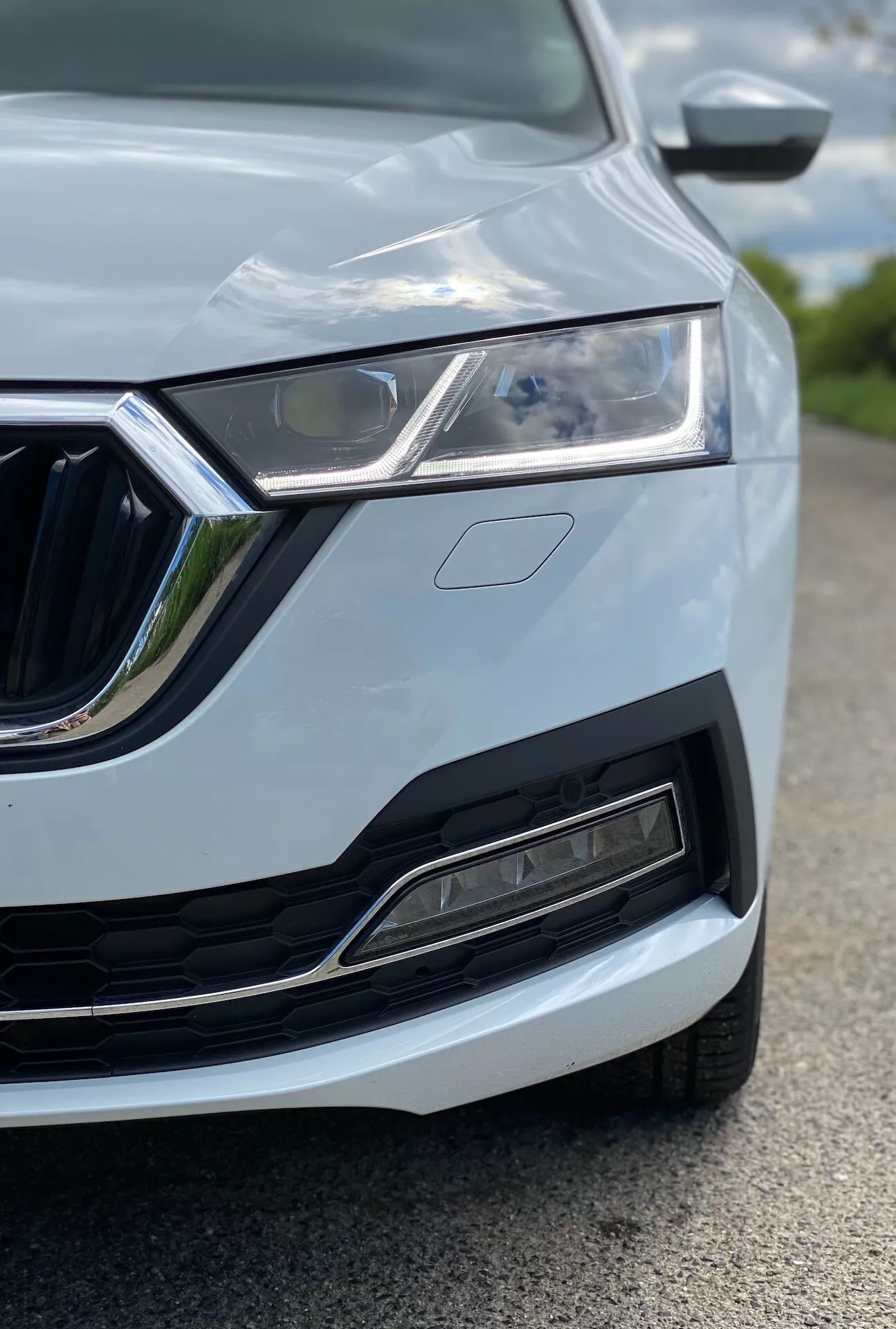 Close-up da parte frontal esquerda de um Skoda Octavia 2020 (octavia-limousine), destacando os faróis LED, a grelha distintiva com detalhes cromados, o para-choques dianteiro moderno com entrada de ar e sensores, design do capô com vinco e o espelho lateral parcialmente visível com piscas integrados. O carro está pintado em uma cor clara, possivelmente branco ou prata, com linhas elegantes da carroçaria. A imagem foi tirada de uma perspectiva baixa, focada no farol e na grelha, com pouca profundidade de campo que desfoca os elementos ao redor.