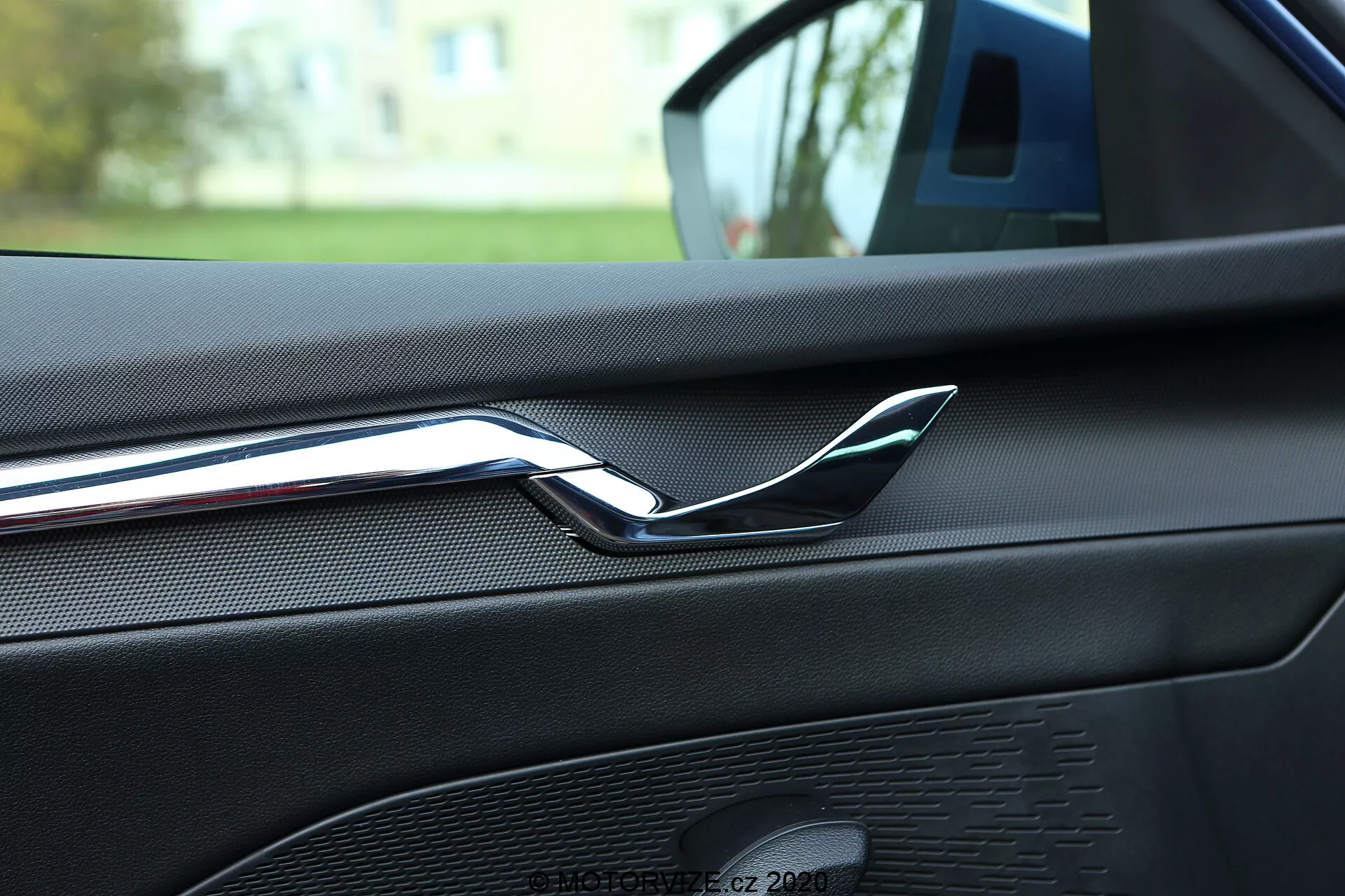 Vista interior em close-up de um Skoda Octavia 2020 focando na parte traseira da porta dianteira do passageiro, destacando a maçaneta de porta cromada reflexiva. O painel da porta apresenta um material em couro sintético ou toque macio texturizado com costuras visíveis, indicativo de um acabamento interior de alta qualidade. Também é visível uma grade de alto-falante como parte do sistema de áudio do carro, com um padrão uniforme e design coordenado em cor. A imagem é tirada do banco traseiro, sugerindo que o carro está estacionado ao ar livre em uma área residencial, com vegetação suavemente desfocada ao fundo.