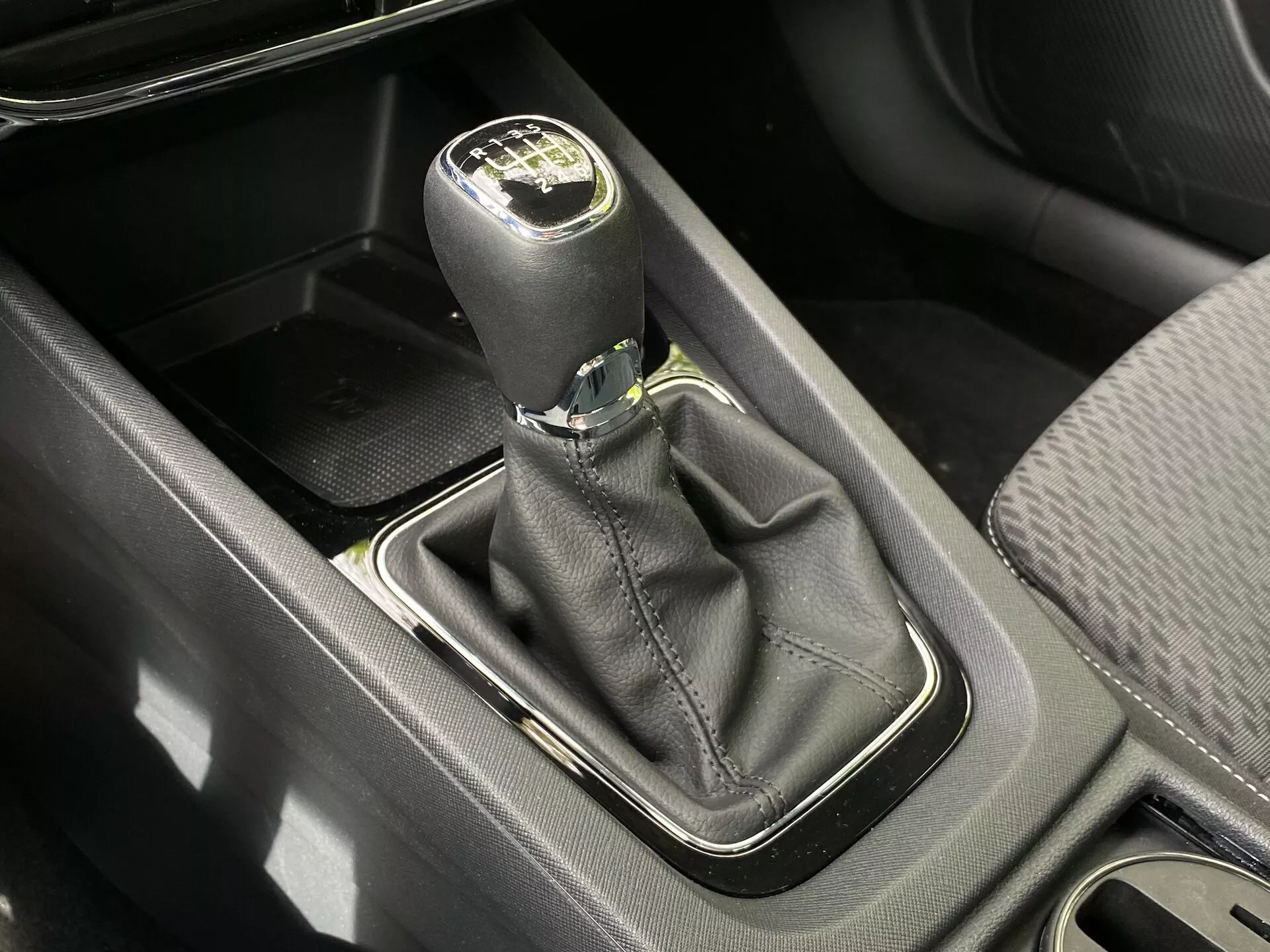 Visão aérea de perto do manípulo de câmbio manual em um Skoda Octavia 2020 (Octavia-Limousine). O manípulo de couro preto com destaque prateado mostra o layout das marchas para seis velocidades mais a ré. A base do manípulo, cercada por uma moldura prateada, está situada em um revestimento de couro preto. Próximos estão um compartimento de armazenamento, console central e porta-copos, todos iluminados pela luz do dia.