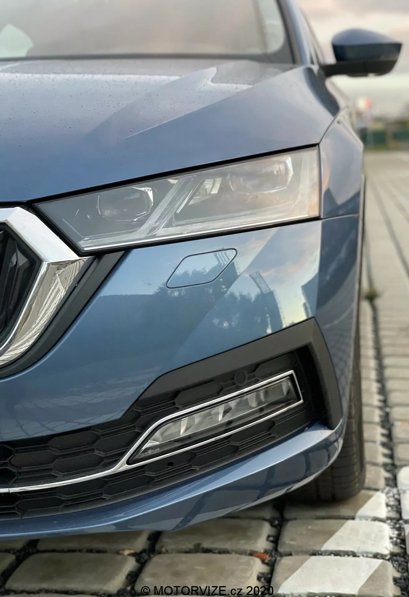 Imagem parcial em close-up de um moderno Skoda Octavia 2020 capturada de um ângulo baixo, focando na seção dianteira esquerda incluindo capô, farol de LED elegante, grade geométrica e para-choque dianteiro com entrada de ar. Pintura azul metálica, elementos de design angulares e características aerodinâmicas destacadas.