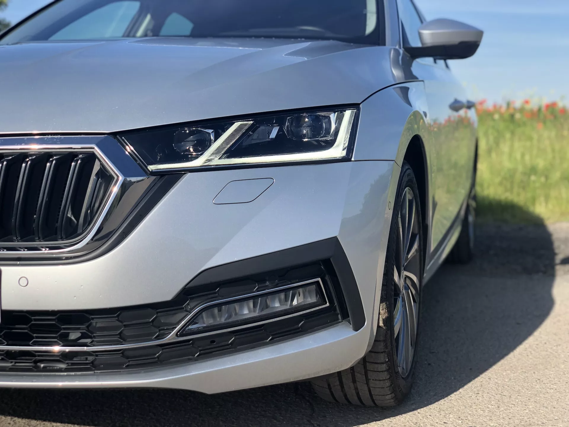 Vista de perto do quarto frontal esquerdo de um Skoda Octavia limousine prateado de 2020, destacando o elegante farol LED, a grade cromada com lâminas verticais, a entrada de ar esculpida e a roda e o para-lama parcialmente visíveis. A foto foi tirada de um ângulo baixo ao nível do solo, focando nos elementos de design moderno e nas características avançadas de tecnologia com um fundo desfocado.