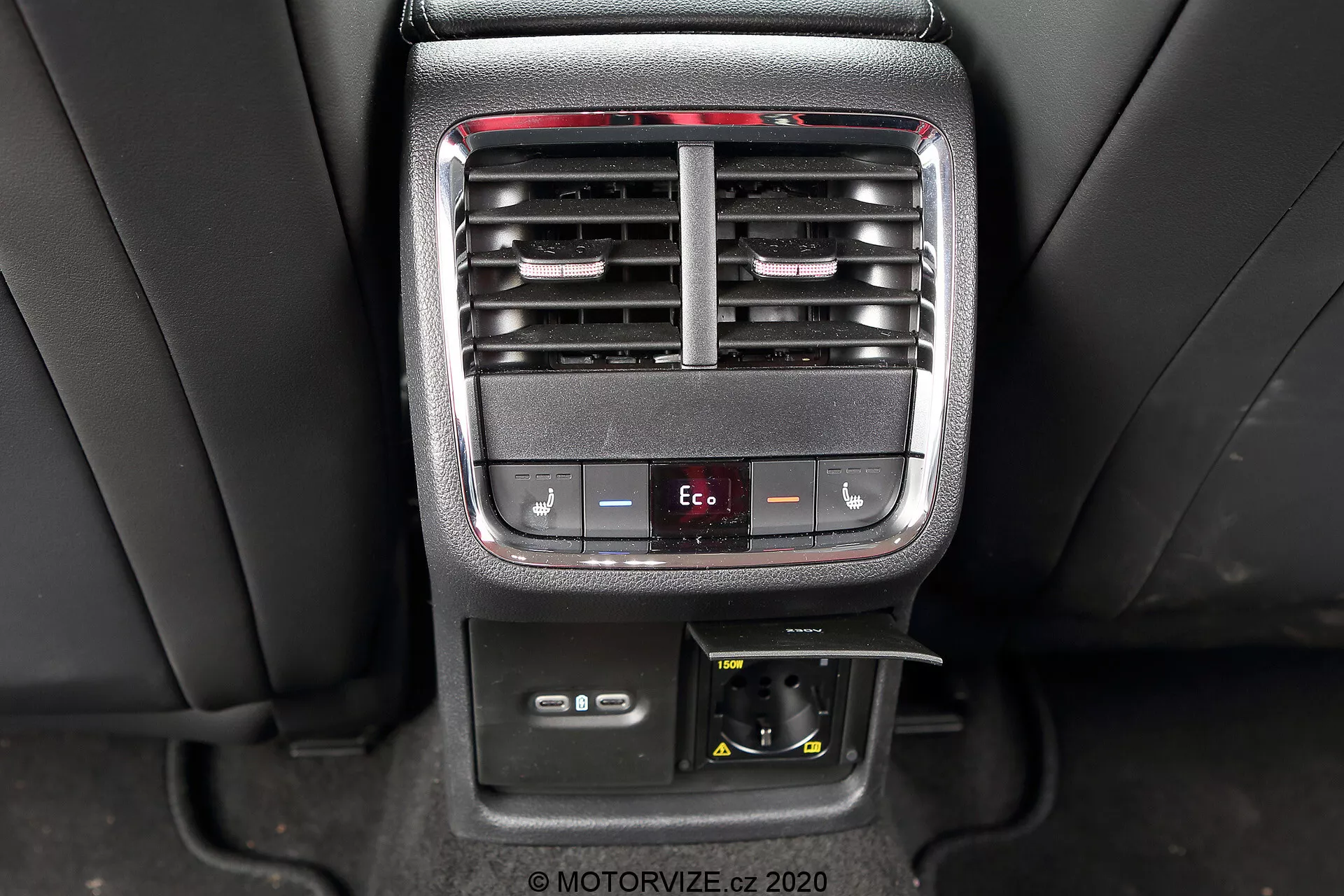 A imagem captura a consola central interior de um Skoda Octavia Limousine 2020, destacando as características de controle de clima e conveniência do passageiro traseiro. A foto oferece uma visão clara das saídas de ar condicionado ajustáveis, botões de controle de temperatura, um botão de modo Eco, uma tomada de corrente de 12V e um compartimento de armazenamento.