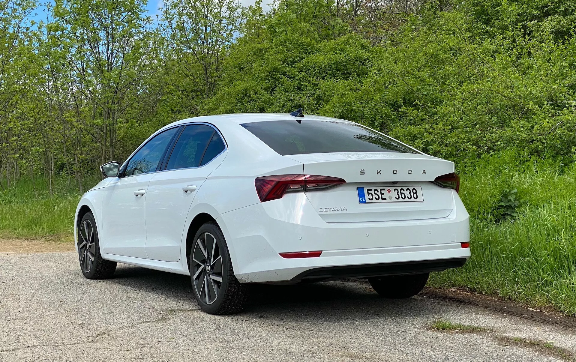 A imagem mostra um sedan Škoda Octavia 2020 branco, capturado de um ângulo traseiro de três quartos, destacando os componentes do lado direito e traseiros do veículo. Detalhes-chave incluem luzes traseiras de LED, logotipo da Škoda e emblema do modelo OCTAVIA, designação TDI, tampa do porta-malas esculpida com spoiler integrado, rodas de liga leve de cinco raios, linha de teto suavemente inclinada, para-choque traseiro com sensores de estacionamento e pintura brilhante e bem conservada. O carro está estacionado em uma tranquila estrada rural ou de parque com vegetação dos dois lados, destacando o ambiente sereno. Esta foto enfatiza o design exterior do carro, as características modernas do sedan e os elementos tecnológicos, como o motor a diesel e os sistemas de assistência ao condutor.