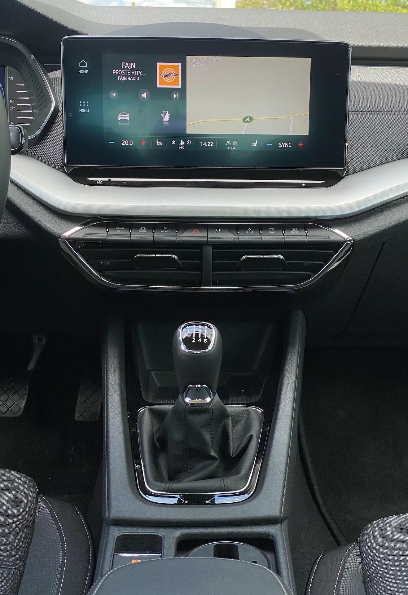 Vista detalhada da console central interior de um Skoda Octavia limusina 2020 a partir da perspectiva do banco traseiro, mostrando uma grande tela sensível ao toque de infotainment com controle de mídia e clima, saídas de ventilação com acentos metálicos e botões de design minimalista, alavanca de câmbio automática estilo manual tradicional com sinais de modo manual, compartimento de armazenamento com portas USB, porta-copos e acabamentos de couro e plástico liso enfatizando conforto moderno e usabilidade centrada no motorista.