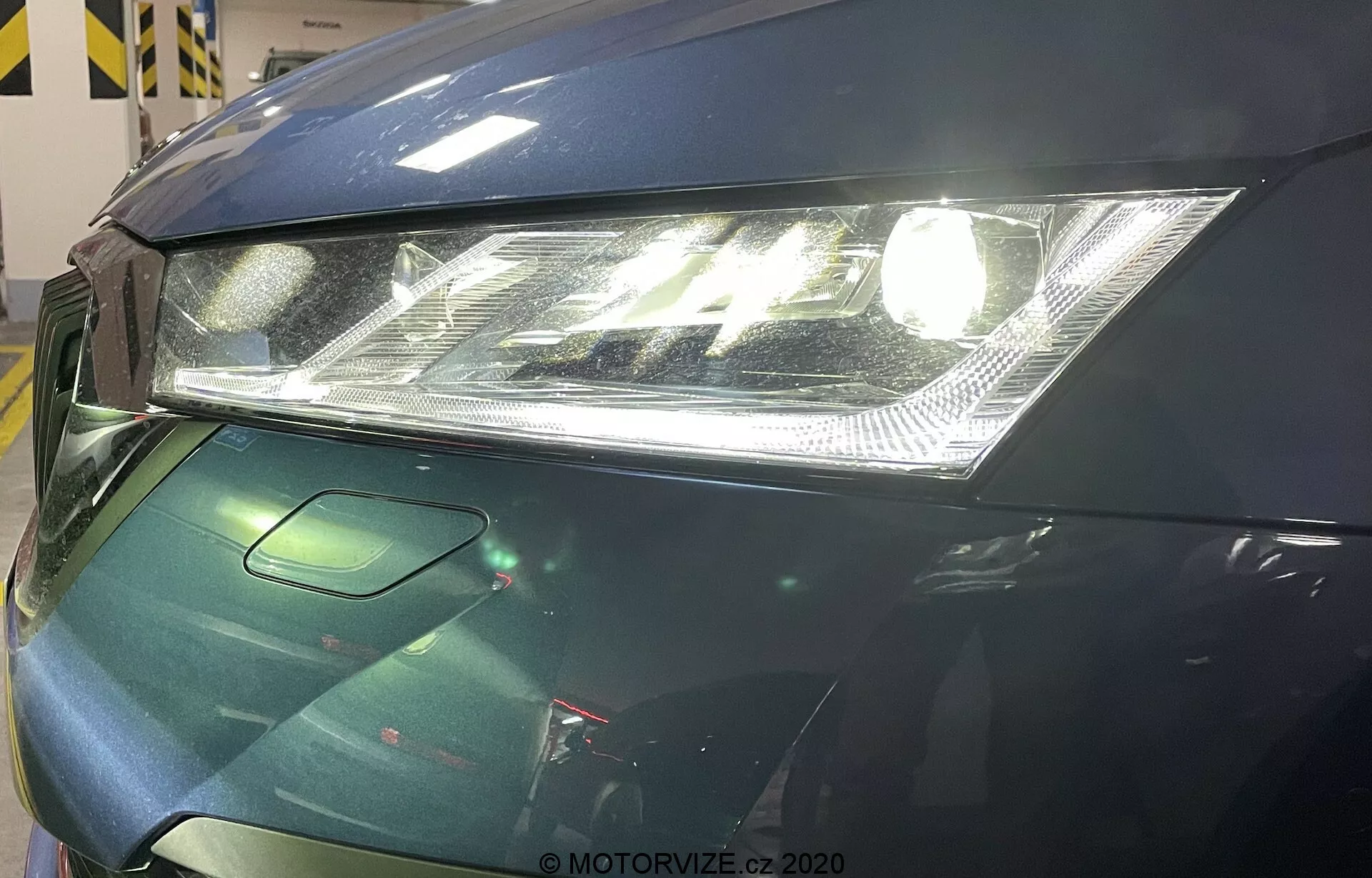 Vista aproximada da área frontal esquerda de um Skoda Octavia 2020. A imagem foca um farol moderno com lente clara e luzes diurnas LED integradas, capô parcialmente visível com design suave e uma pequena seção da grade provavelmente incorporando sensores de assistência ao motorista. O carro apresenta um acabamento brilhante na cor verde escuro, sugerindo medidas avançadas de proteção da pintura.