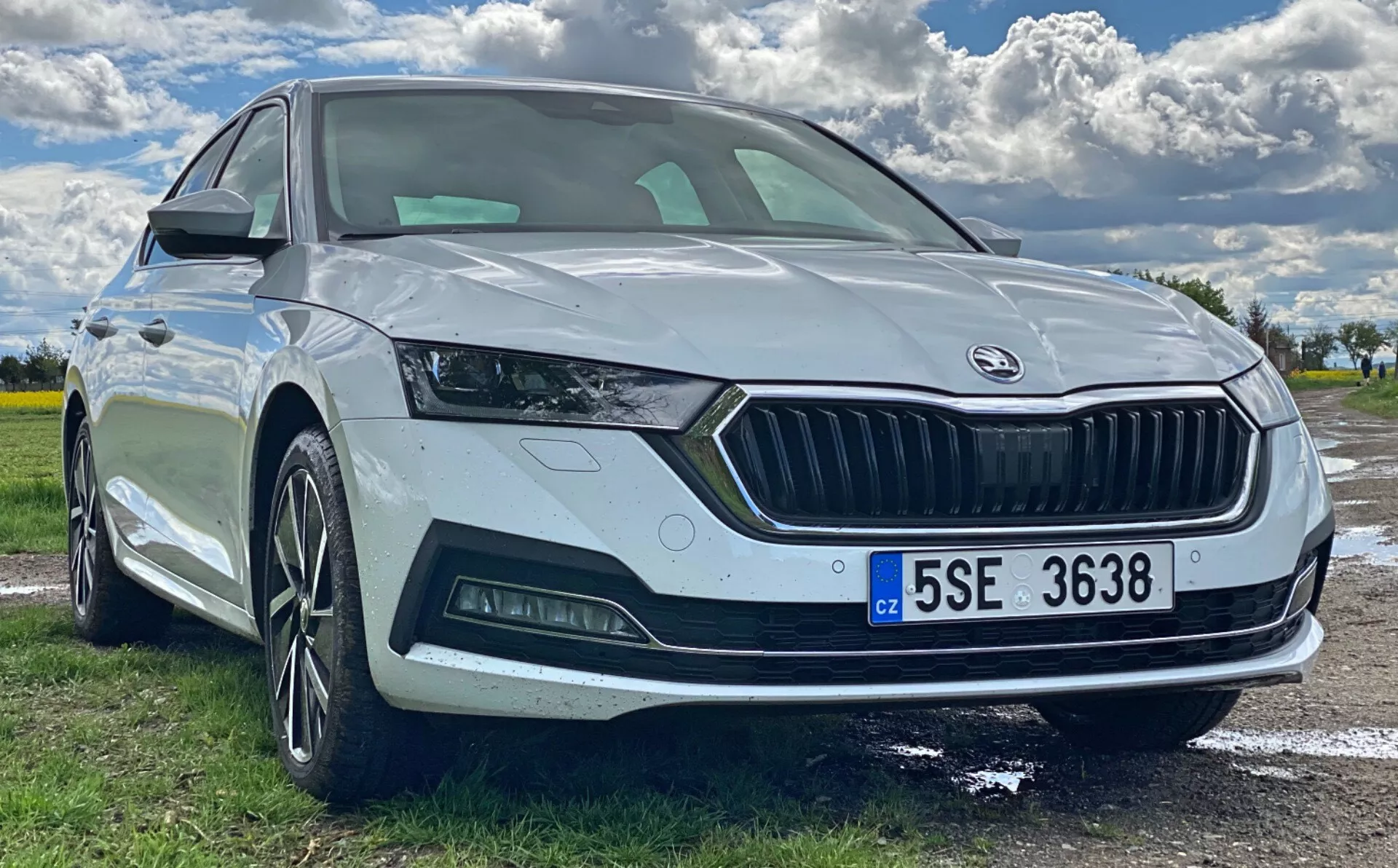 Imagem em ângulo baixo do quarto dianteiro de um Škoda Octavia 2020 branco estacionado ao ar livre, destacando a região frontal, design angular dos faróis com luzes diurnas de LED, grade larga com logotipo Škoda, sensor de estacionamento, para-choque dianteiro esportivo com faróis de neblina, rodas de liga leve de múltiplos raios e indicador de direção integrado no retrovisor lateral. O para-brisa panorâmico e a placa de licença estilo europeu também são visíveis.